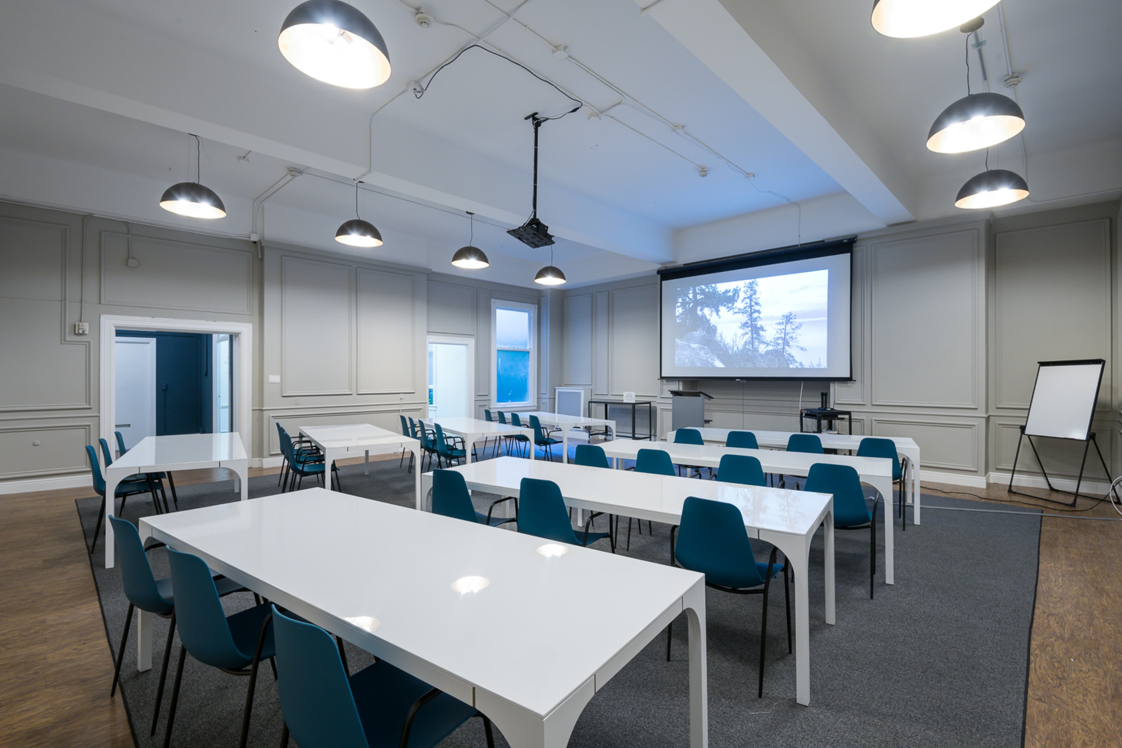 A spacious conference room features white tables arranged in a U-shape, blue chairs, and a projector screen at the front.