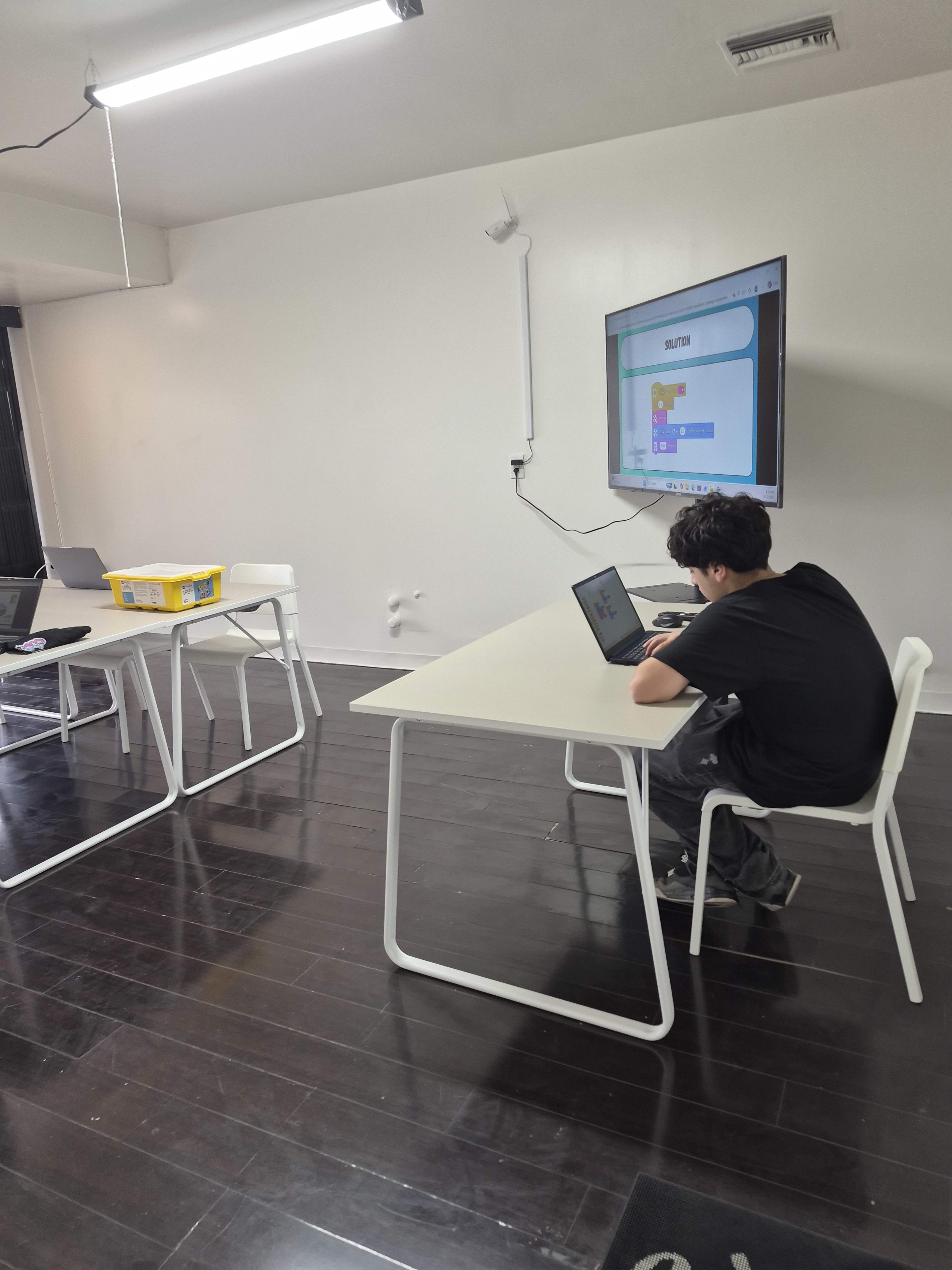 A student sits at a table, using a laptop in a mostly empty classroom with a large screen displaying content on the wall.
