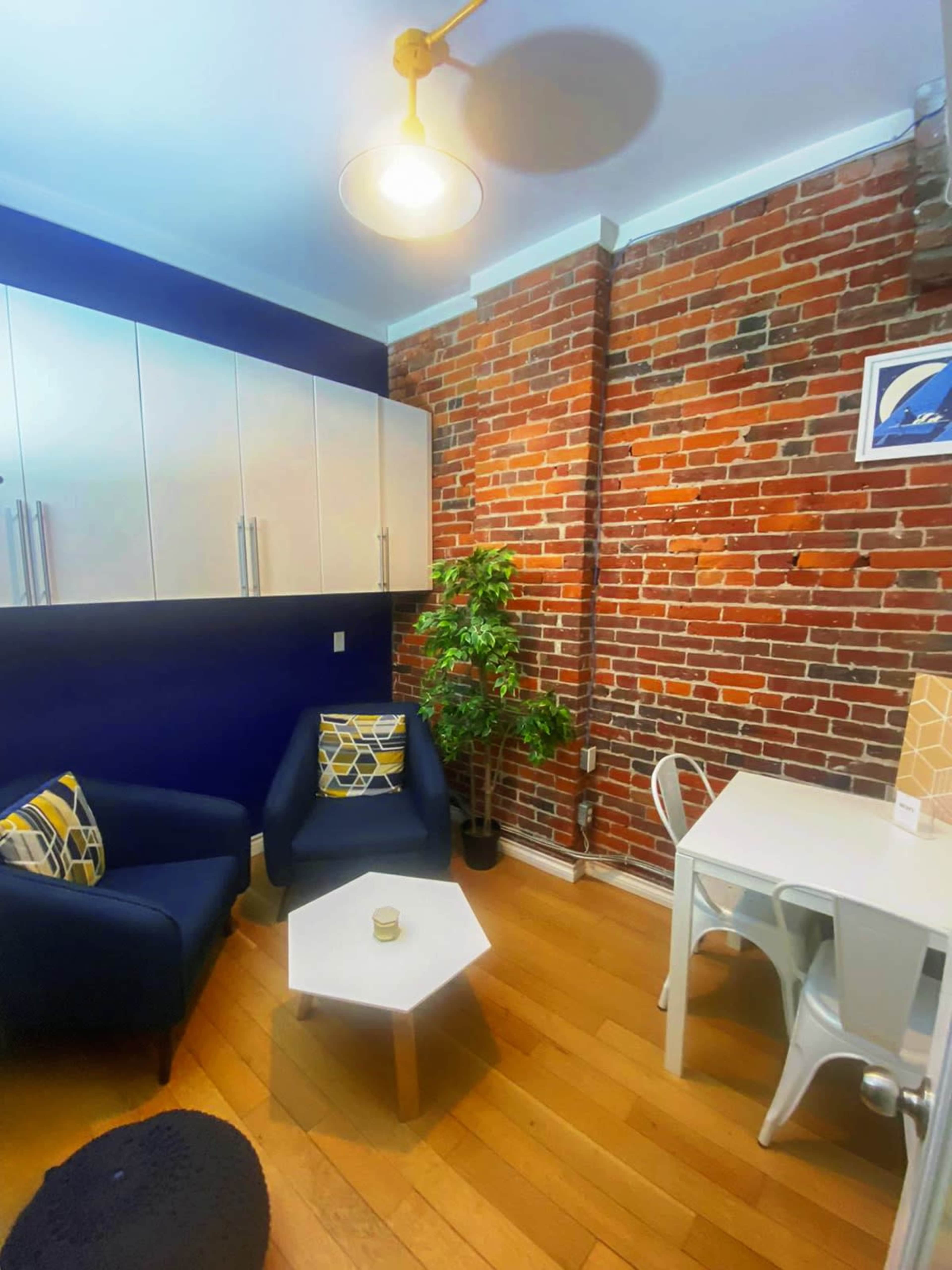 A small living space featuring a brick wall, a blue sofa, a small coffee table, a white dining table with chairs, and a potted plant.