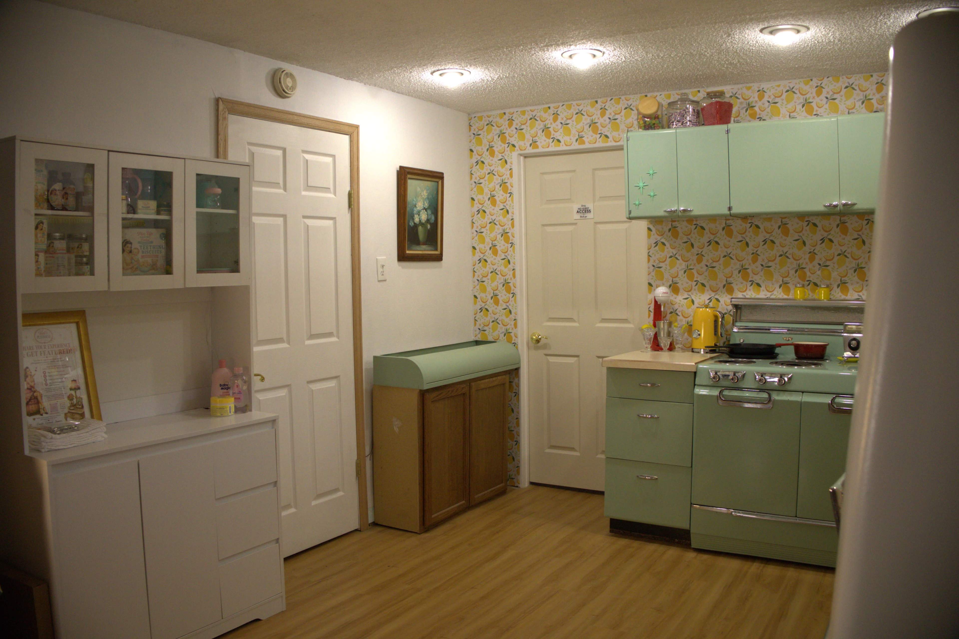 The image shows a retro-styled kitchen featuring mint green appliances, floral wallpaper, and white cabinetry.