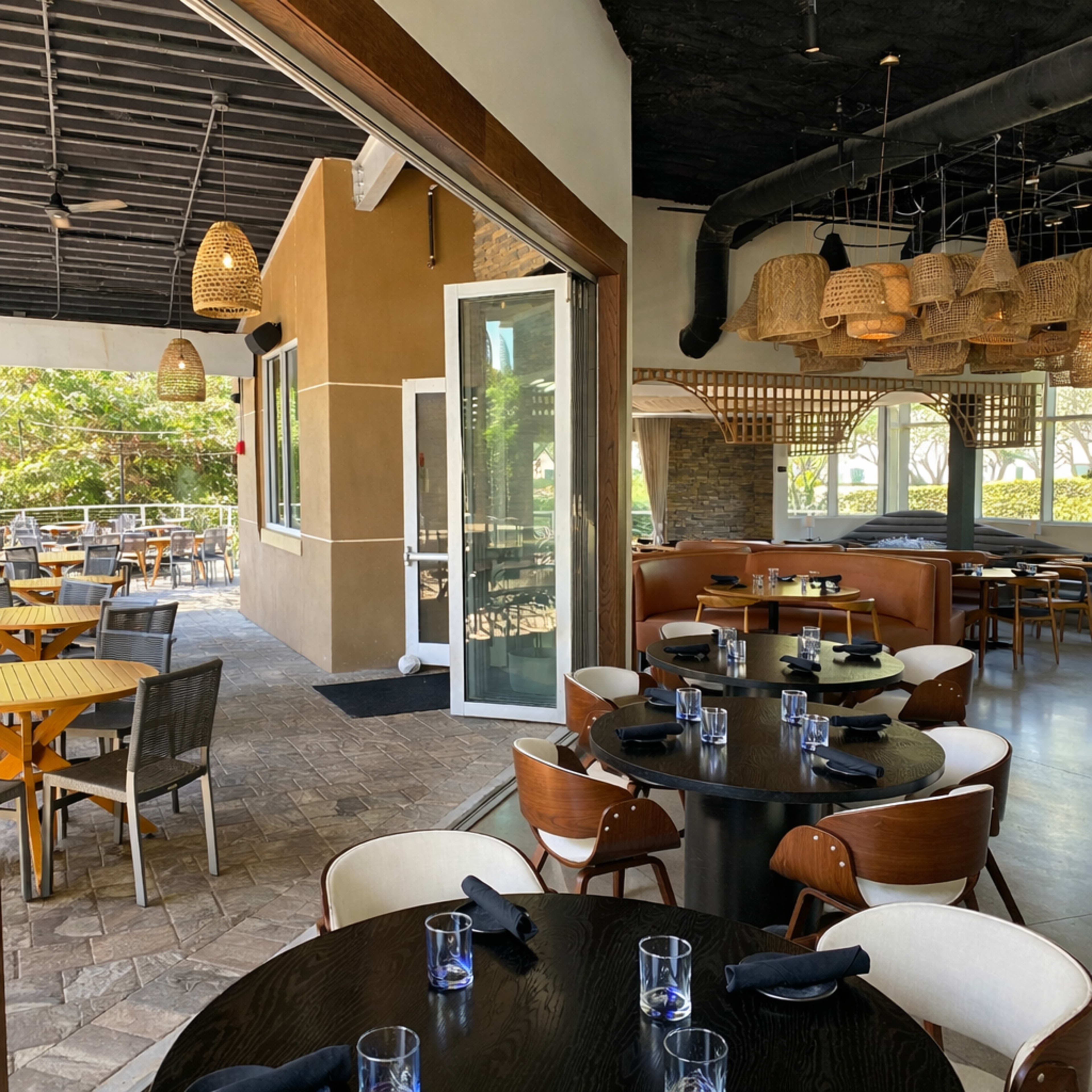 The image shows an upscale restaurant with a modern interior featuring round and square tables set for dining, and a view of an outdoor seating area through a large entrance.