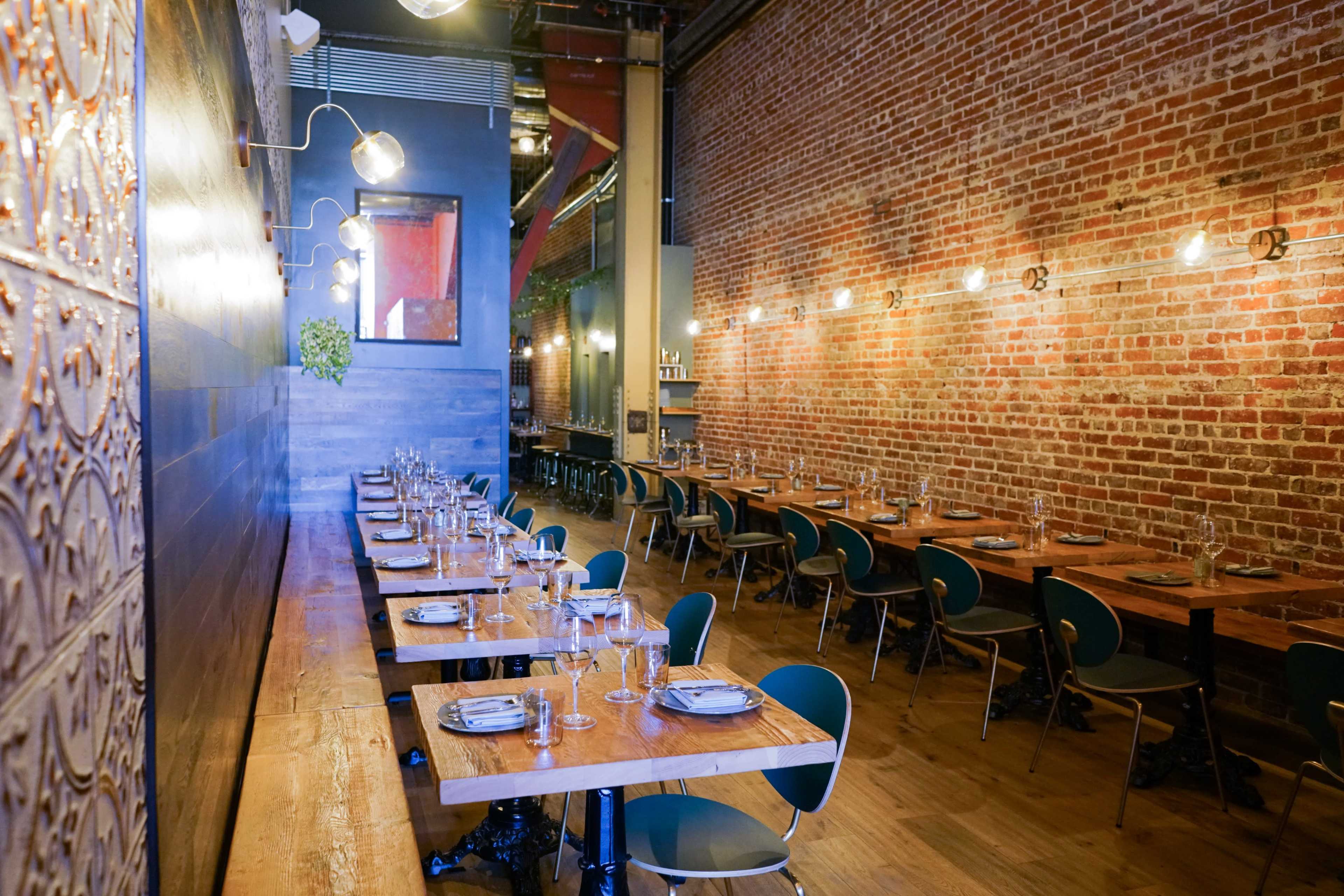 The image shows a spacious restaurant interior with brick walls, wooden tables set for dining, and green chairs arranged in rows.