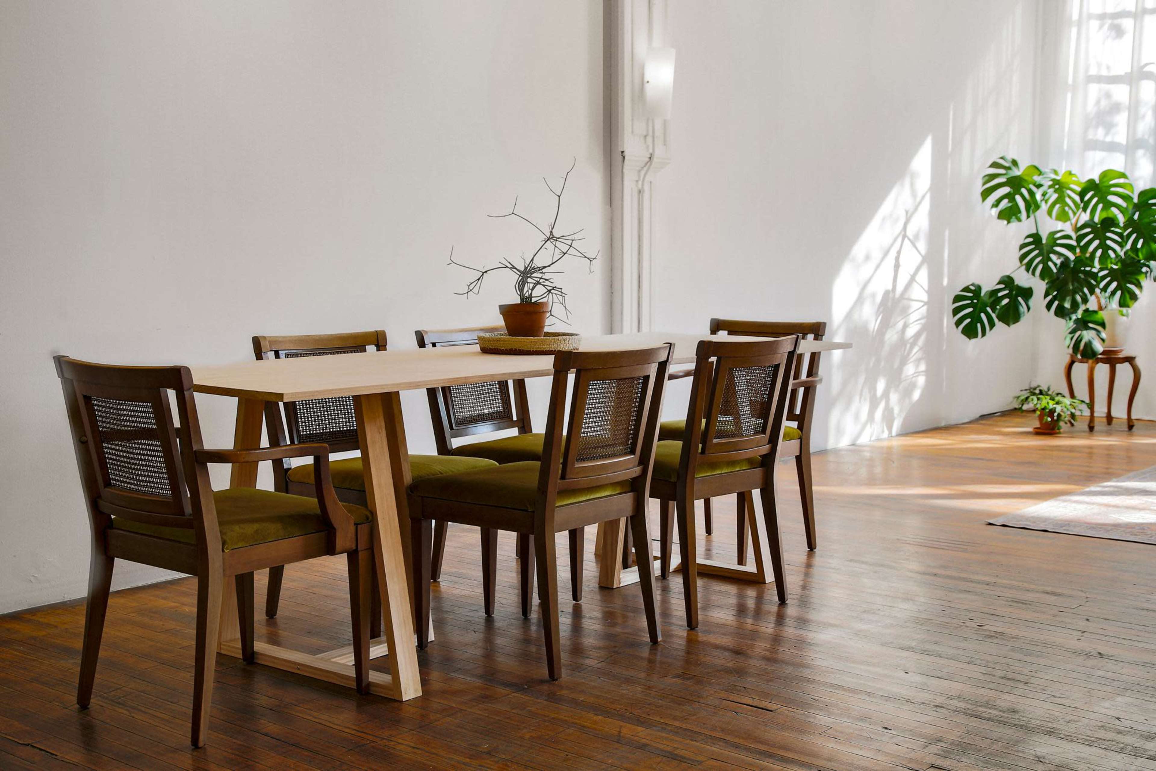 A wooden dining table with six chairs sits in a bright room featuring large windows and a potted plant.