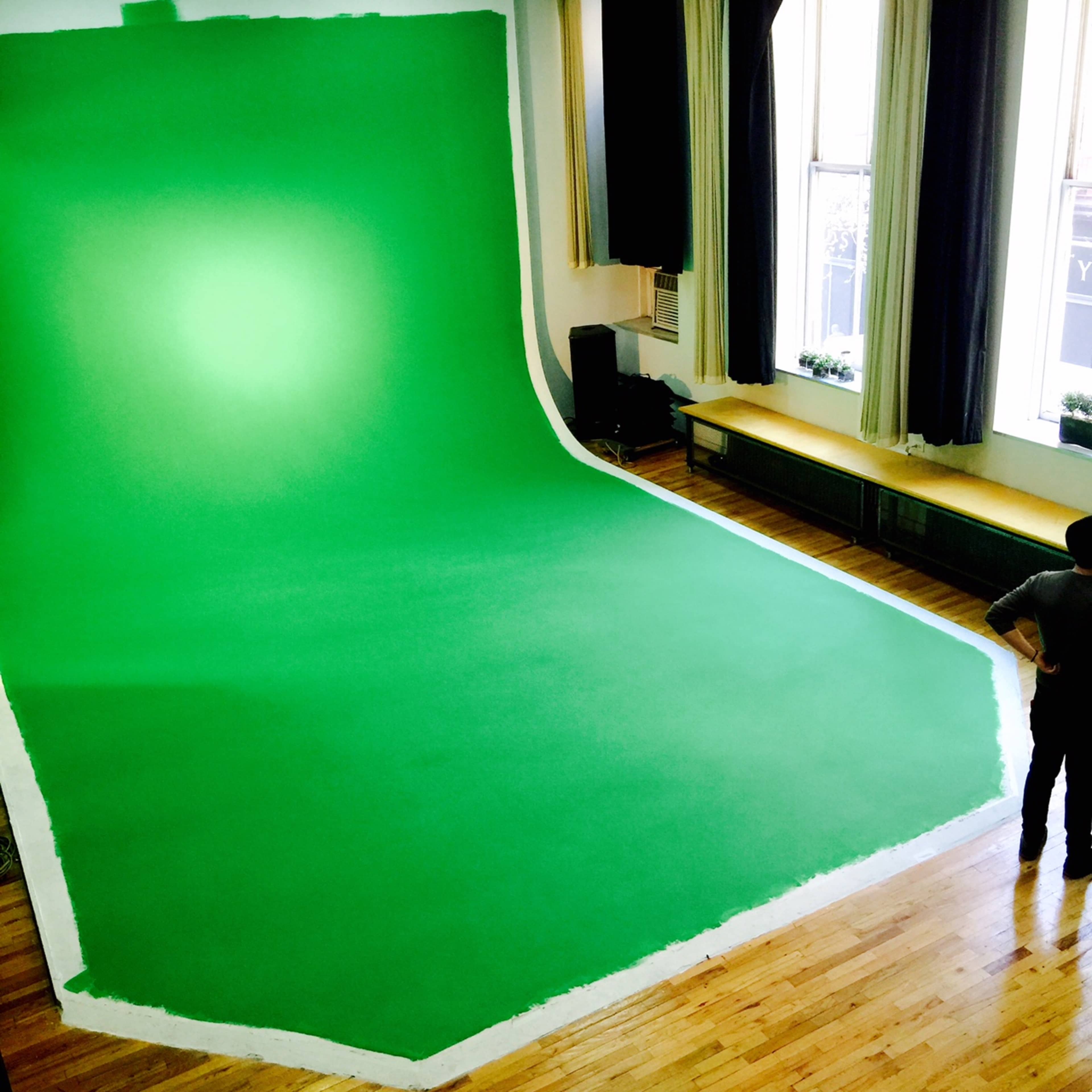 A large green backdrop is set up in a room with wooden floors and windows, while a person stands nearby.