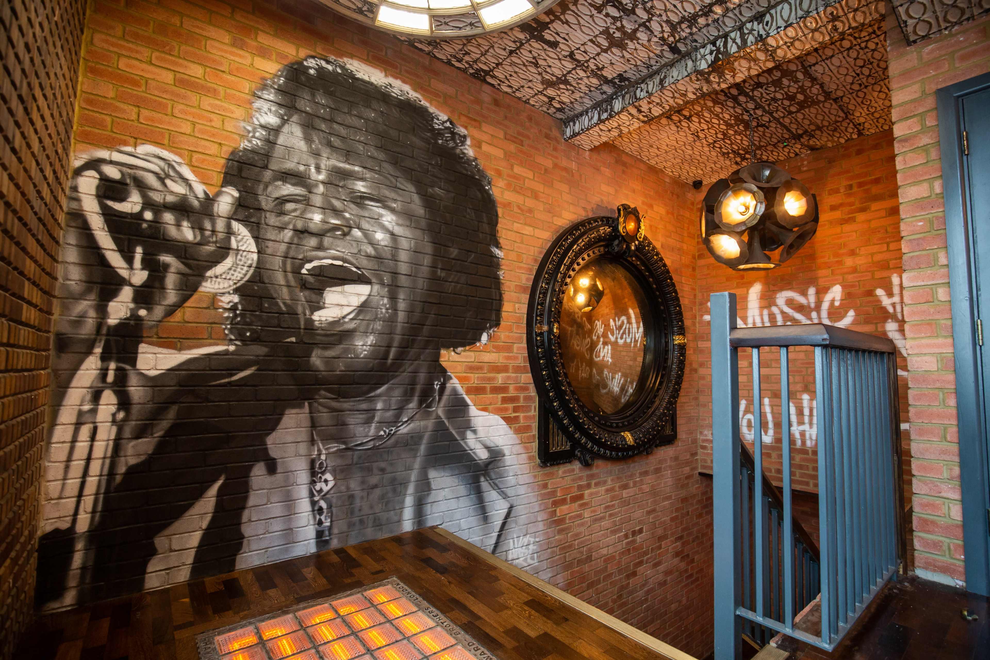 A large mural of a singer adorns a brick wall in a stairwell featuring industrial-style lighting and a circular mirror.