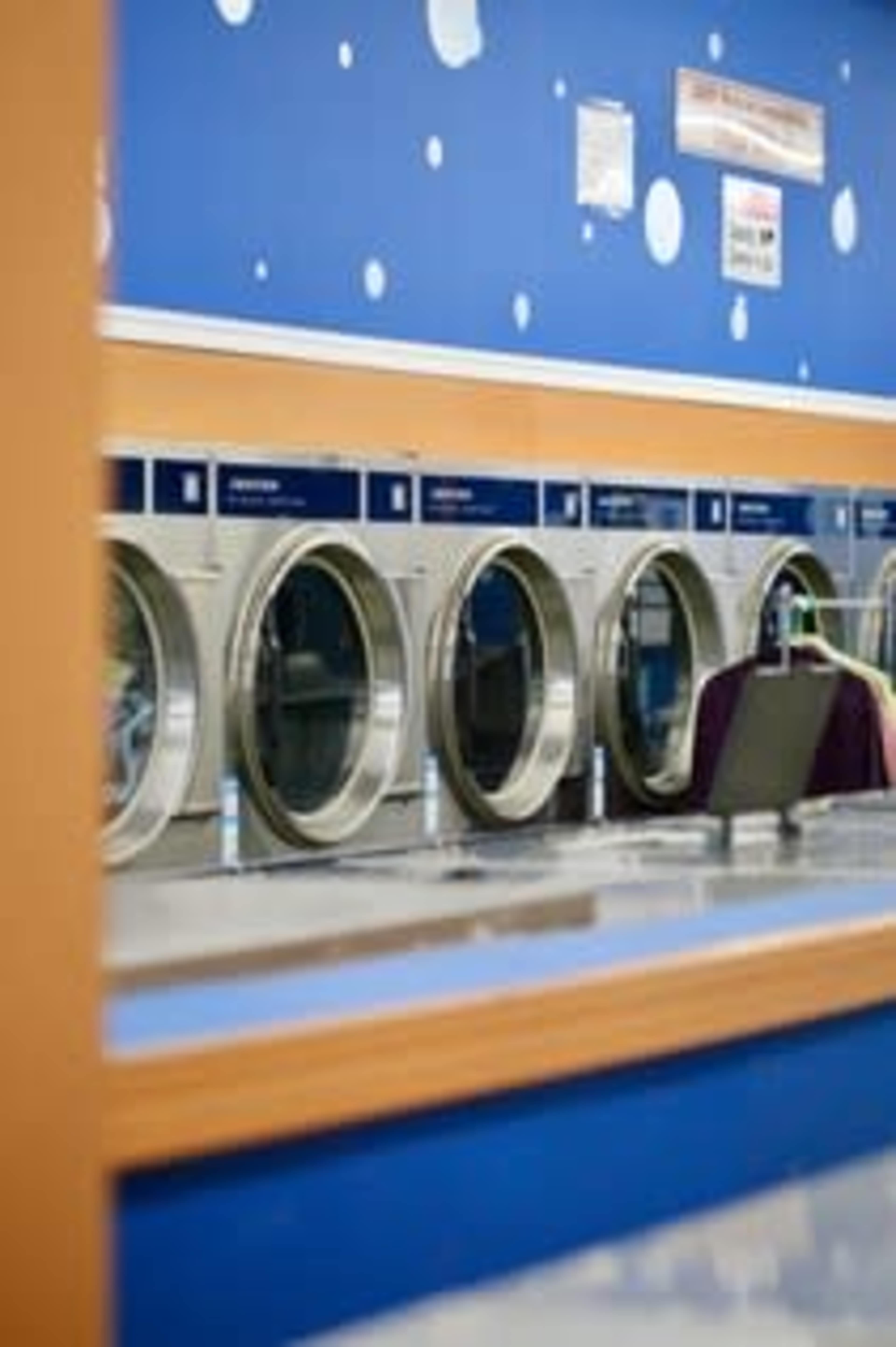 The image shows a row of stainless steel washing machines in a laundromat with a blue wall decorated with white dots.