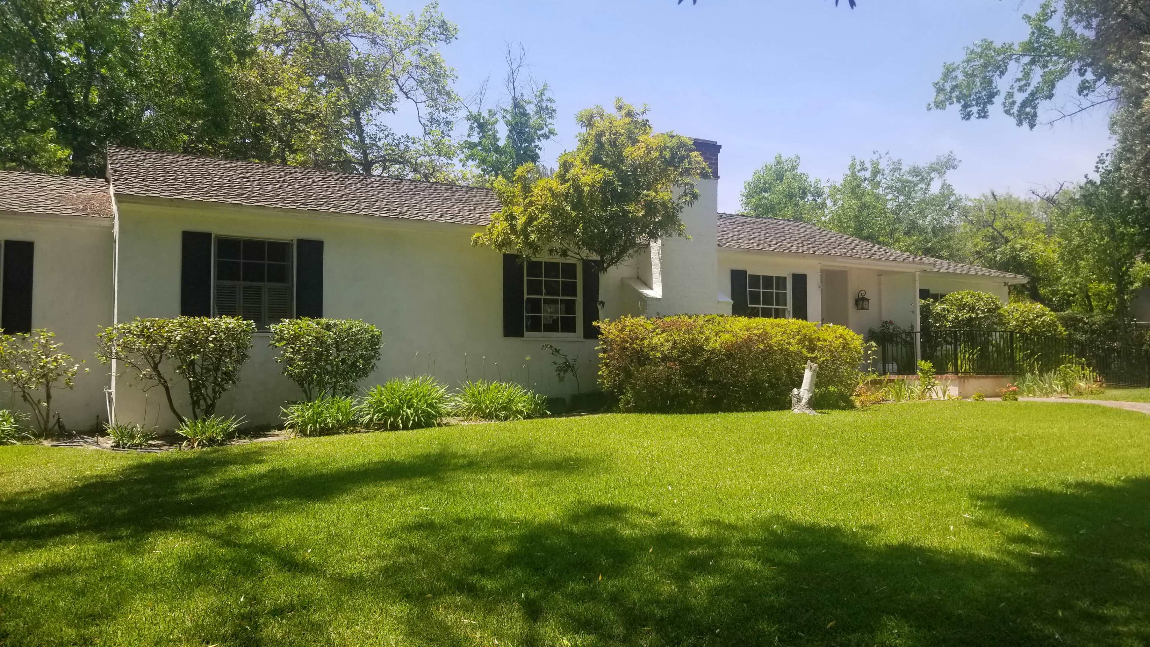 A single-story white house with a dark roof and black shutters sits amid green grass and shrubs in a well-landscaped yard.
