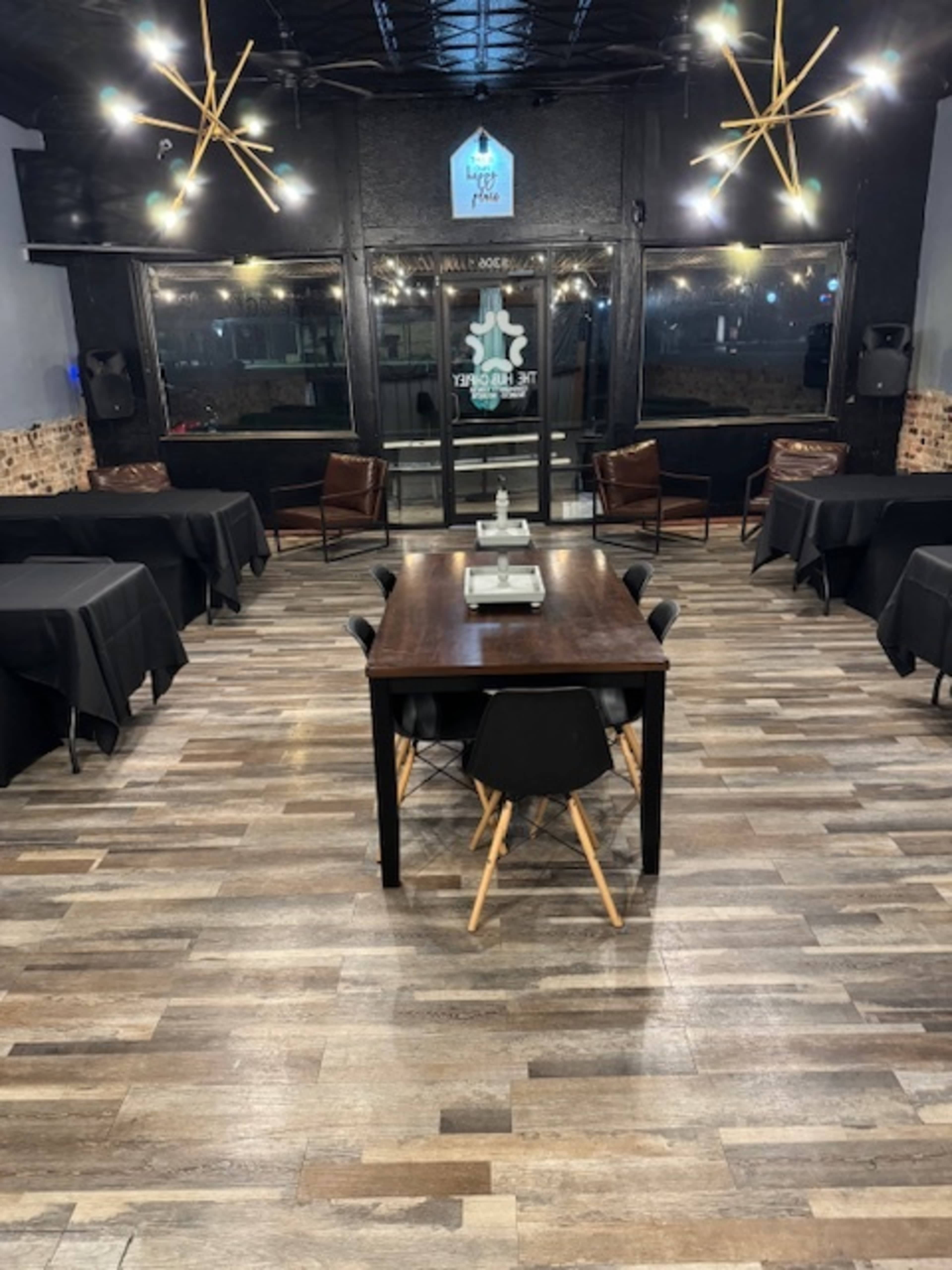 The image shows a restaurant interior with several tables covered in black tablecloths, a large central wooden table, and modern light fixtures hanging from the ceiling.