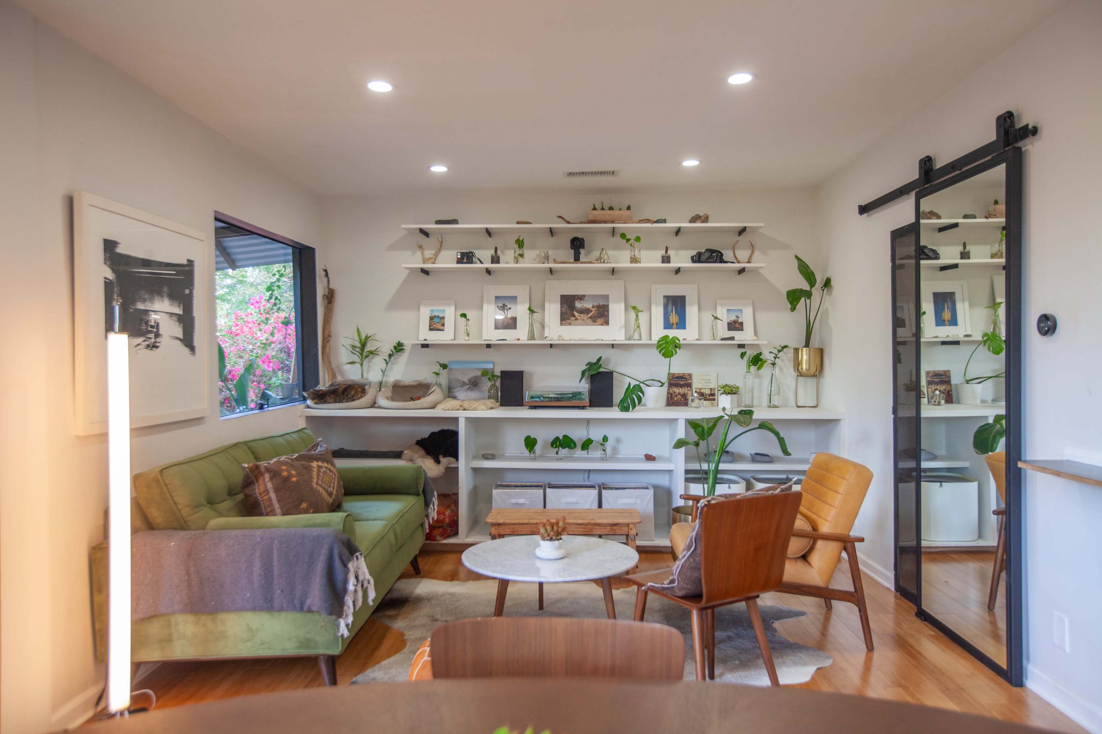 A modern living room features a green sofa, a circular coffee table, and shelves displaying decorative items and plants, with a sliding glass door at one side.