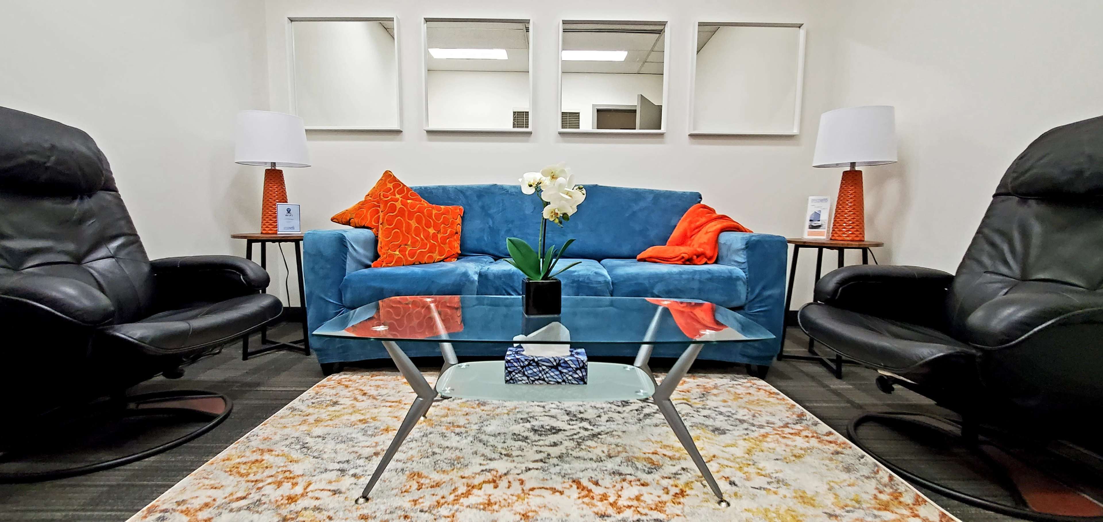 A modern seating area with a blue sofa, two black armchairs, a glass coffee table, and decorative cushions, surrounded by mirrors on the wall.