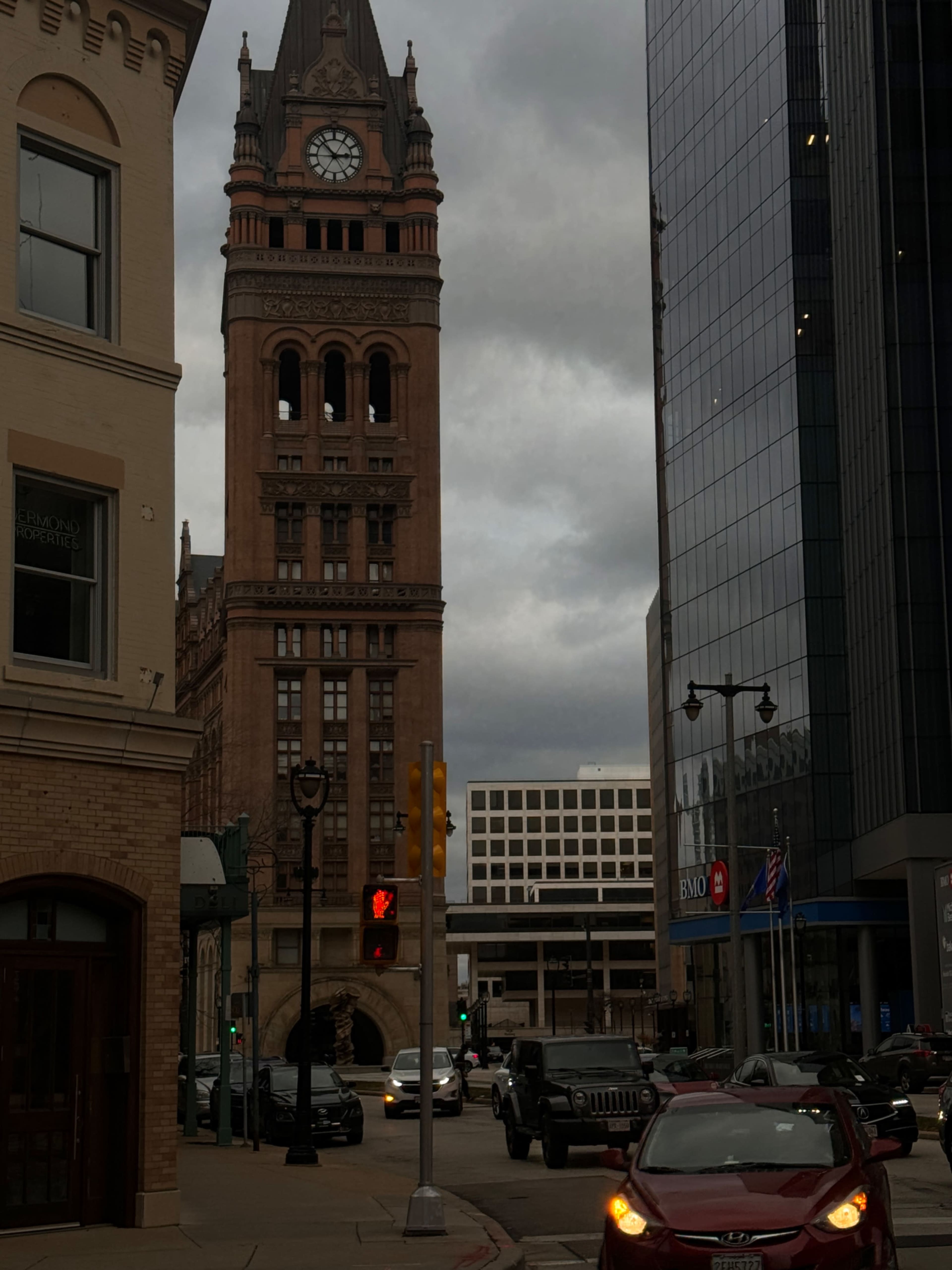 A tall clock tower stands at the end of a city street, framed by modern buildings and overcast skies.