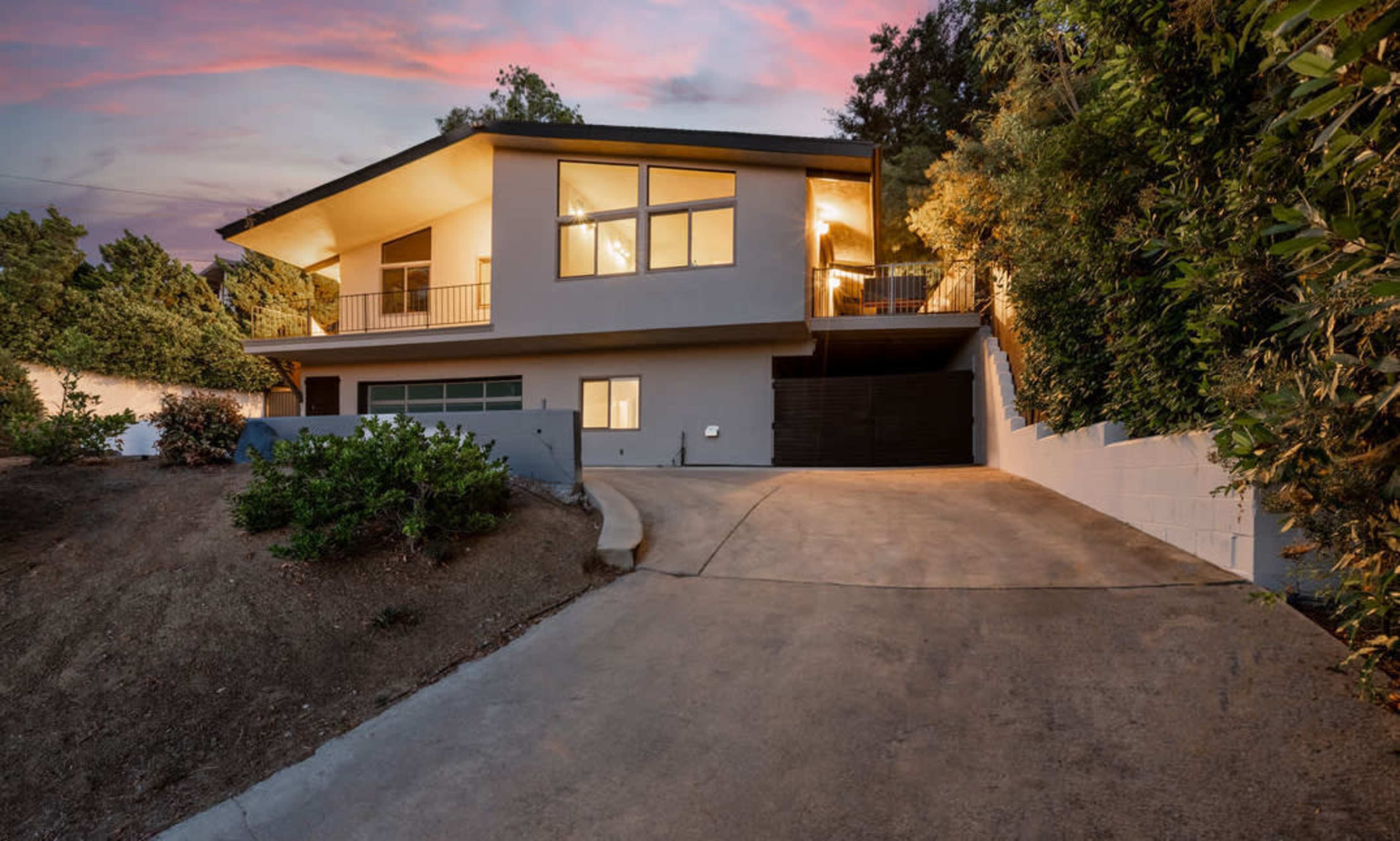 A modern house with large windows is situated on a sloped driveway surrounded by greenery at dusk.