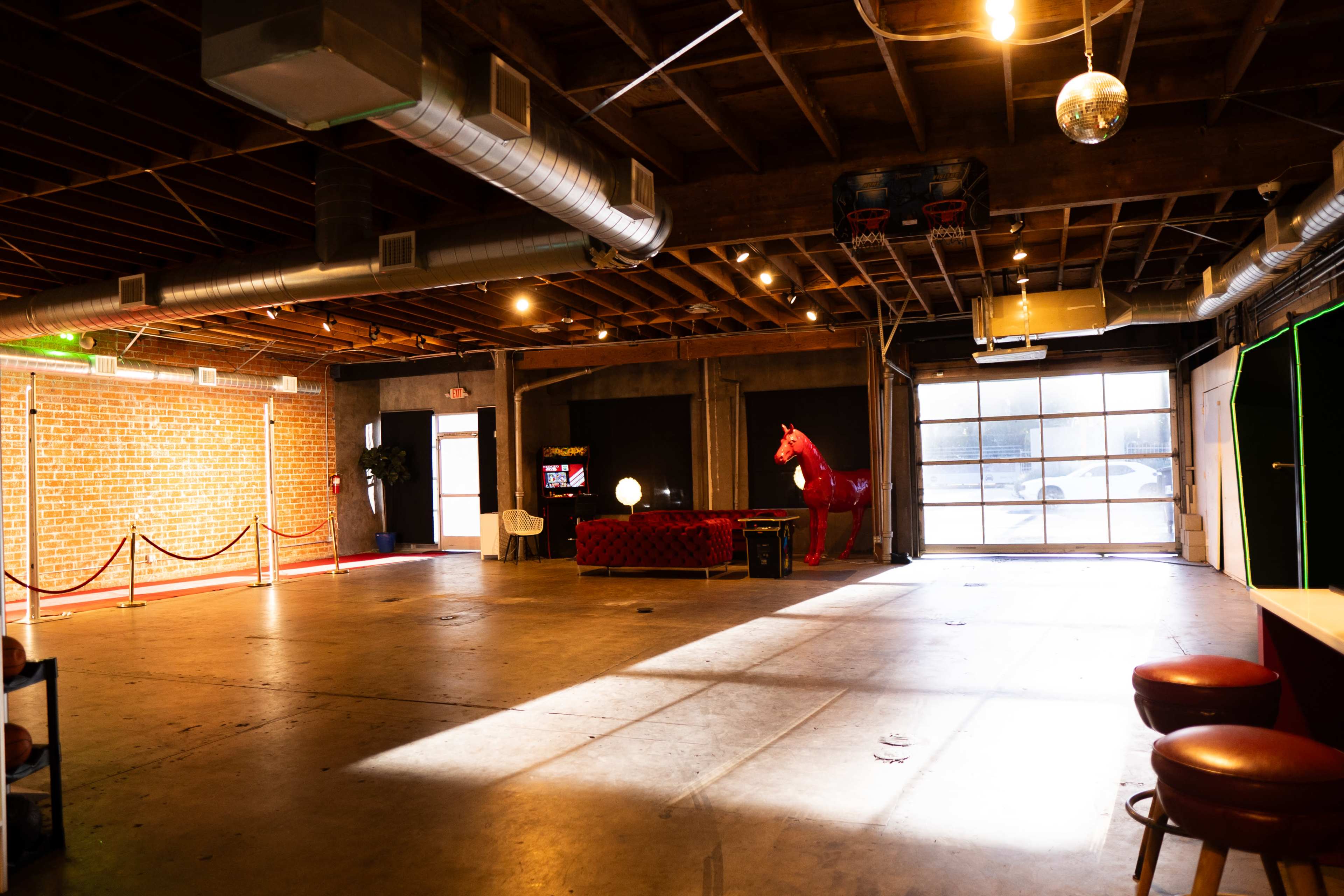 The image shows an empty industrial space with exposed wooden beams and brick walls, featuring a red horse statue, a table, and lighting elements.