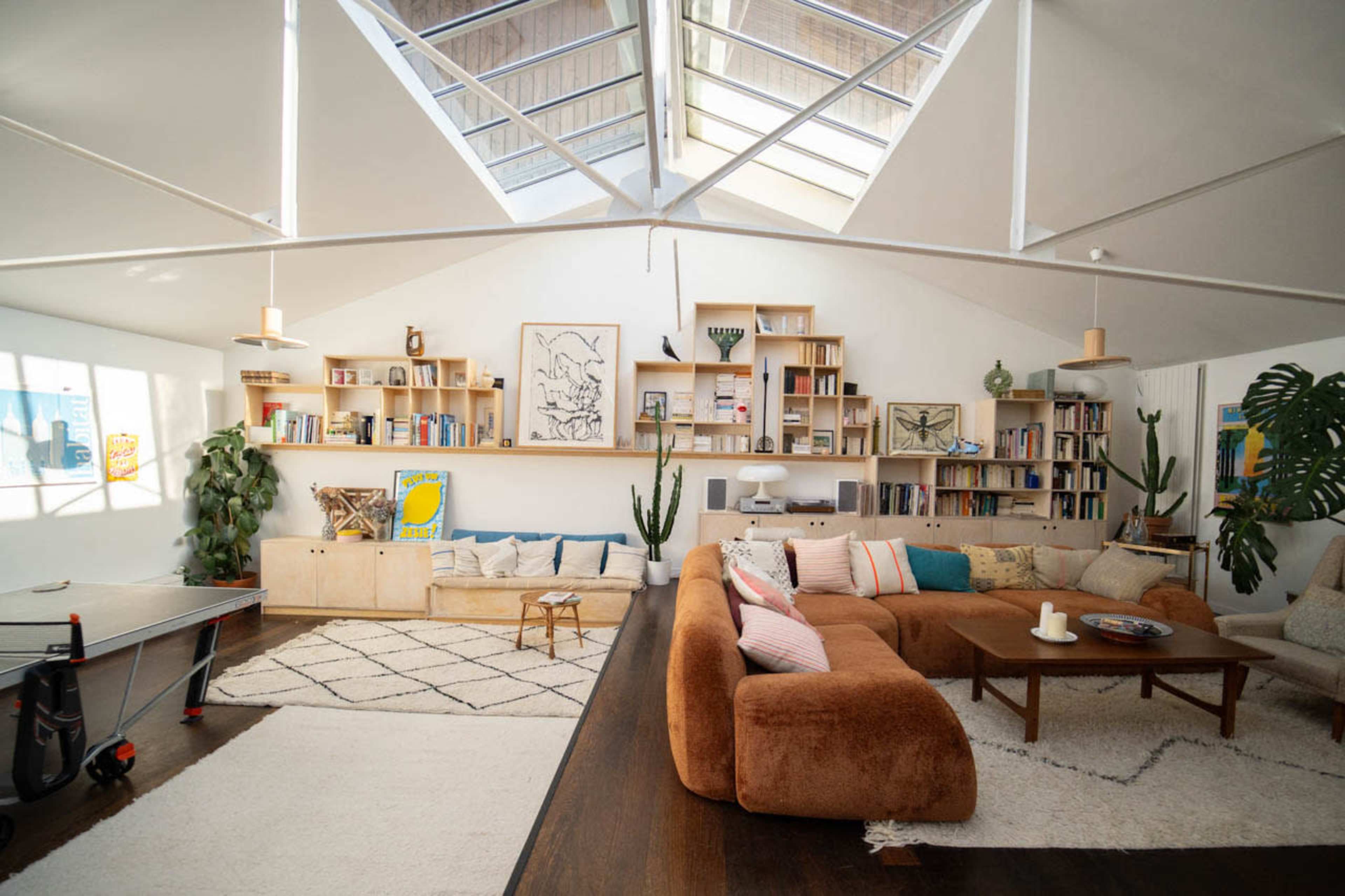 The image shows a spacious, well-lit living room featuring a large sofa, bookshelves, indoor plants, and a table tennis setup.