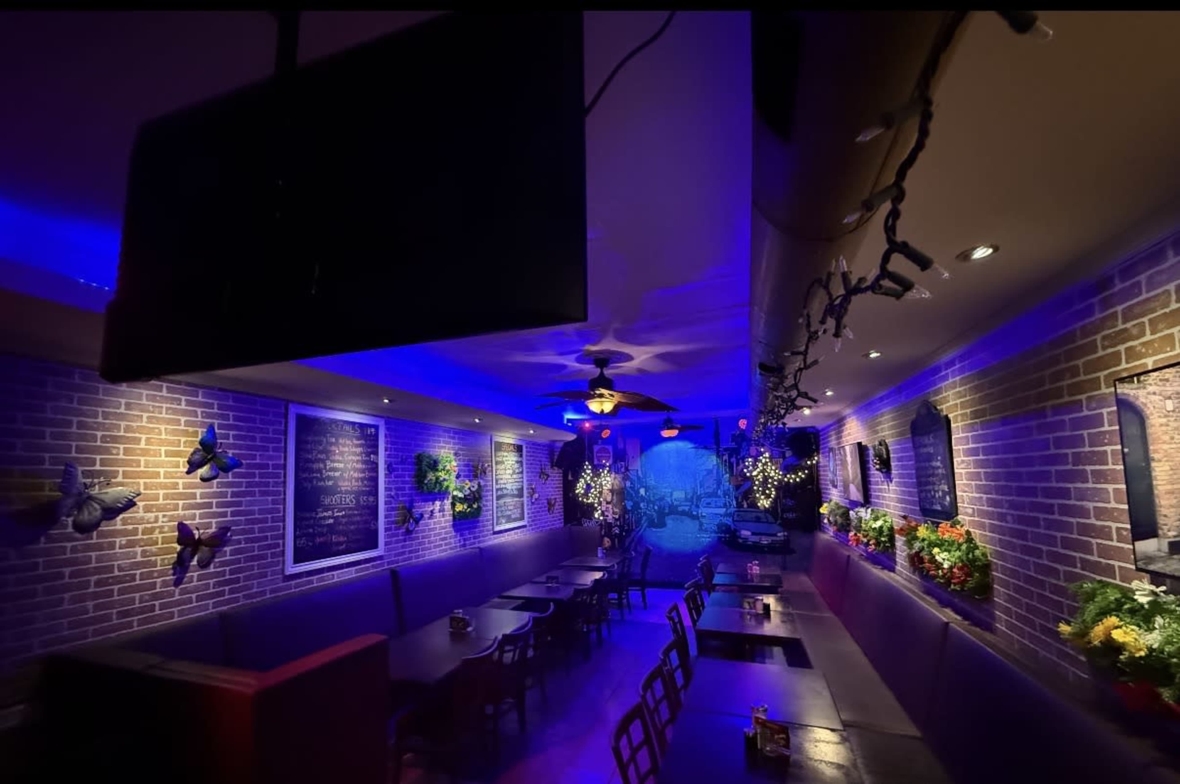 The image shows a dimly lit restaurant interior with brick walls, a long table lined with chairs, and decorative plants, illuminated by blue and purple lighting.