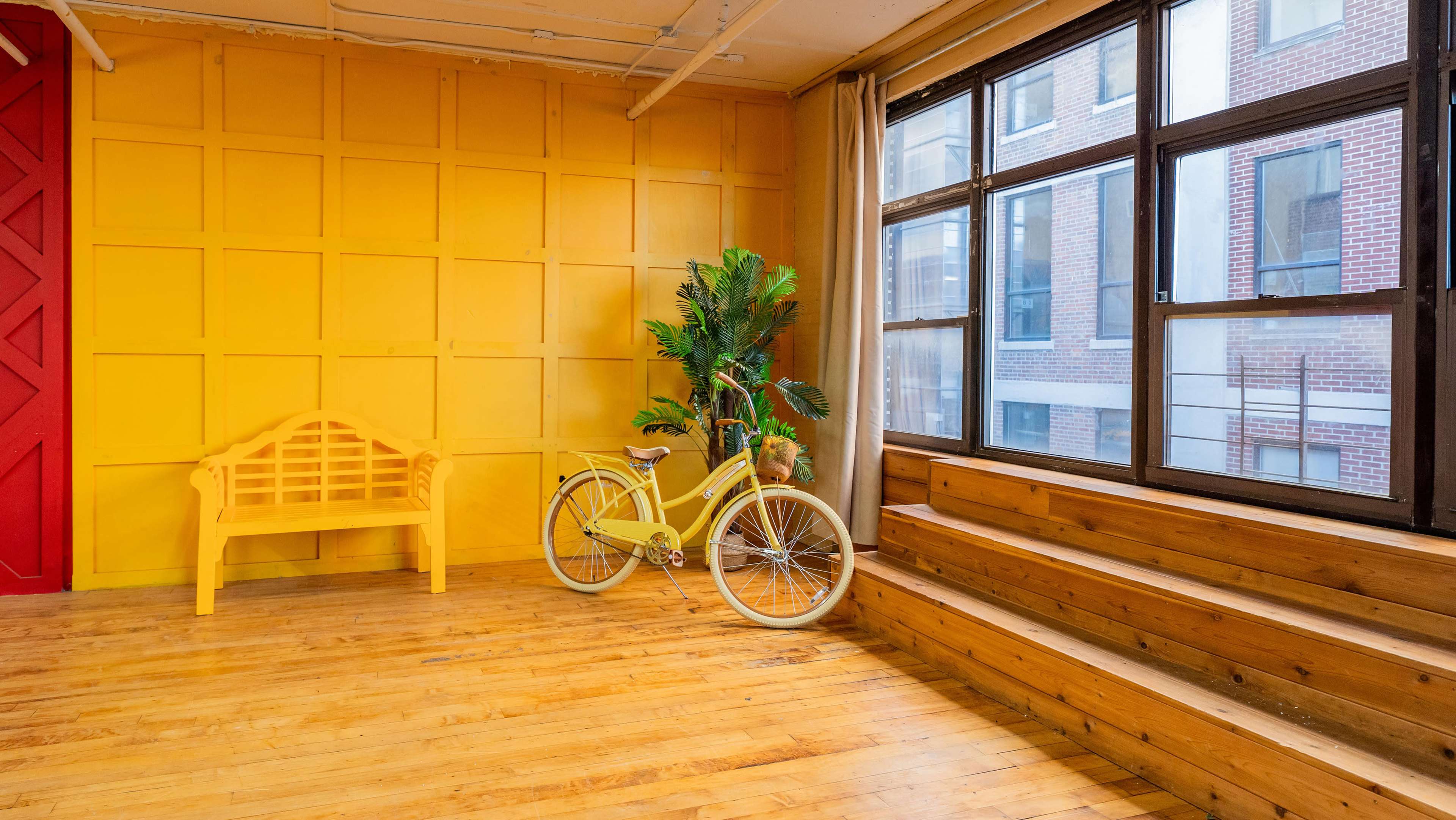 A brightly colored room with yellow walls, a wooden floor, a yellow bicycle, and a yellow bench beside a large window.
