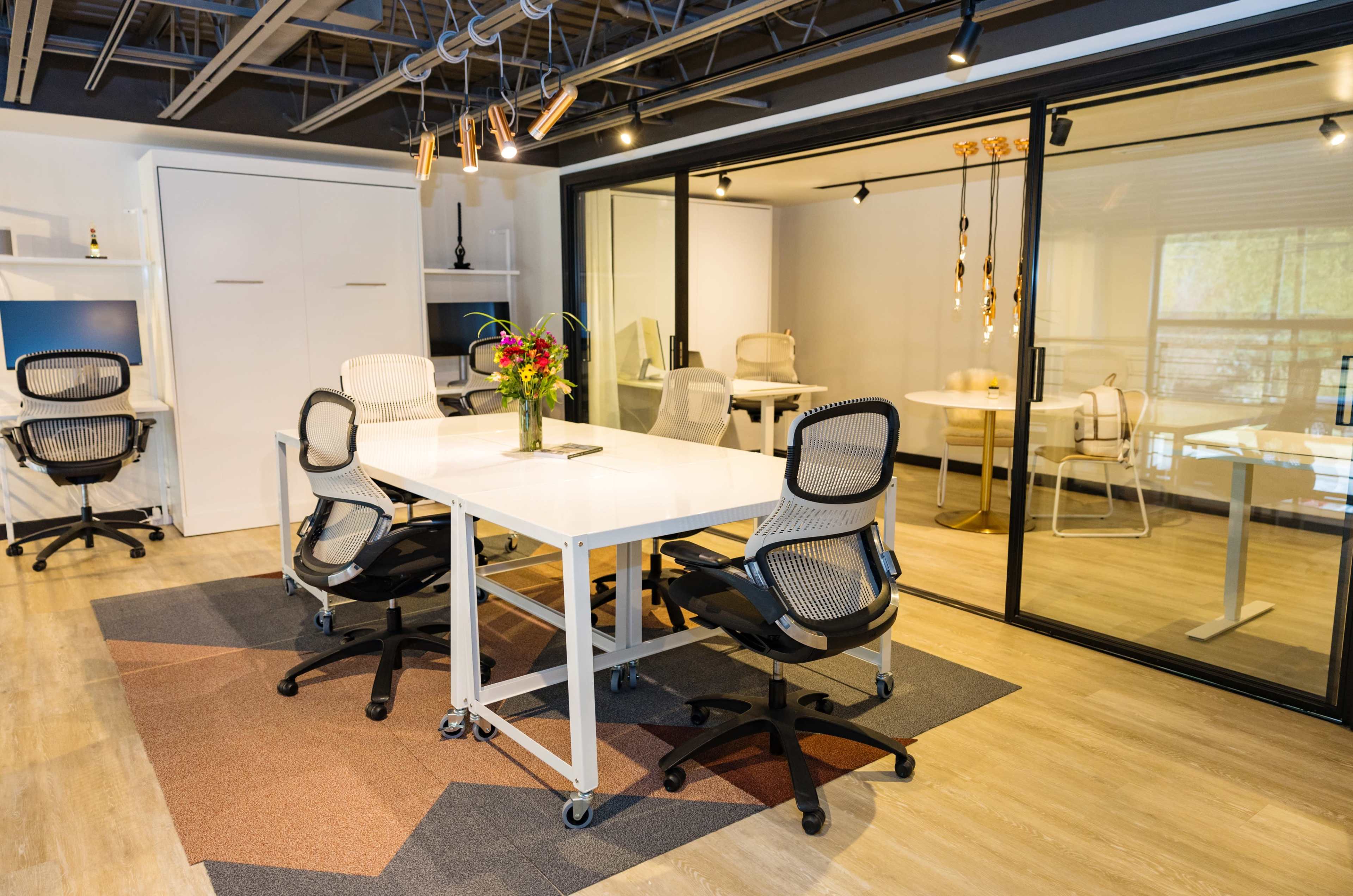 The scene shows a modern office space with a central white table surrounded by rolling office chairs, and a glass-walled conference room in the background.