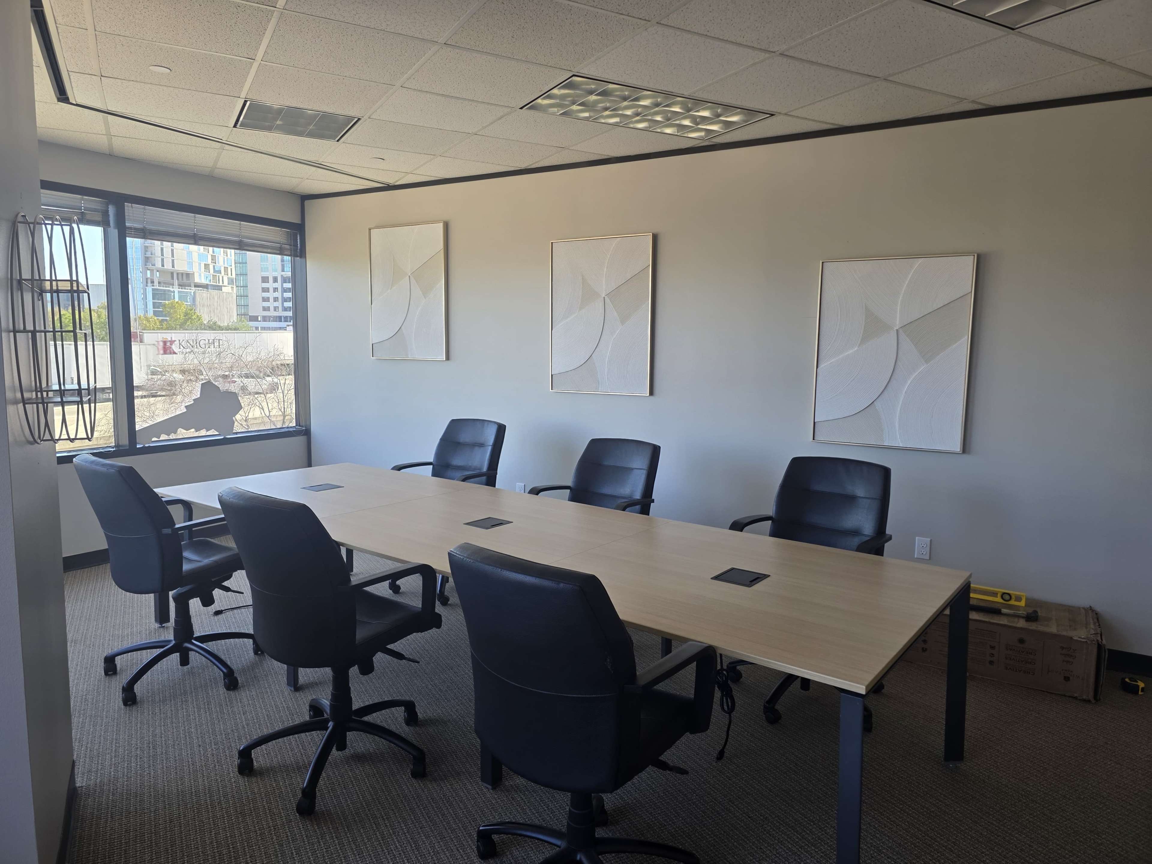 The image shows a modern conference room with a long table and six black chairs arranged around it, featuring three abstract wall art pieces and a large window.