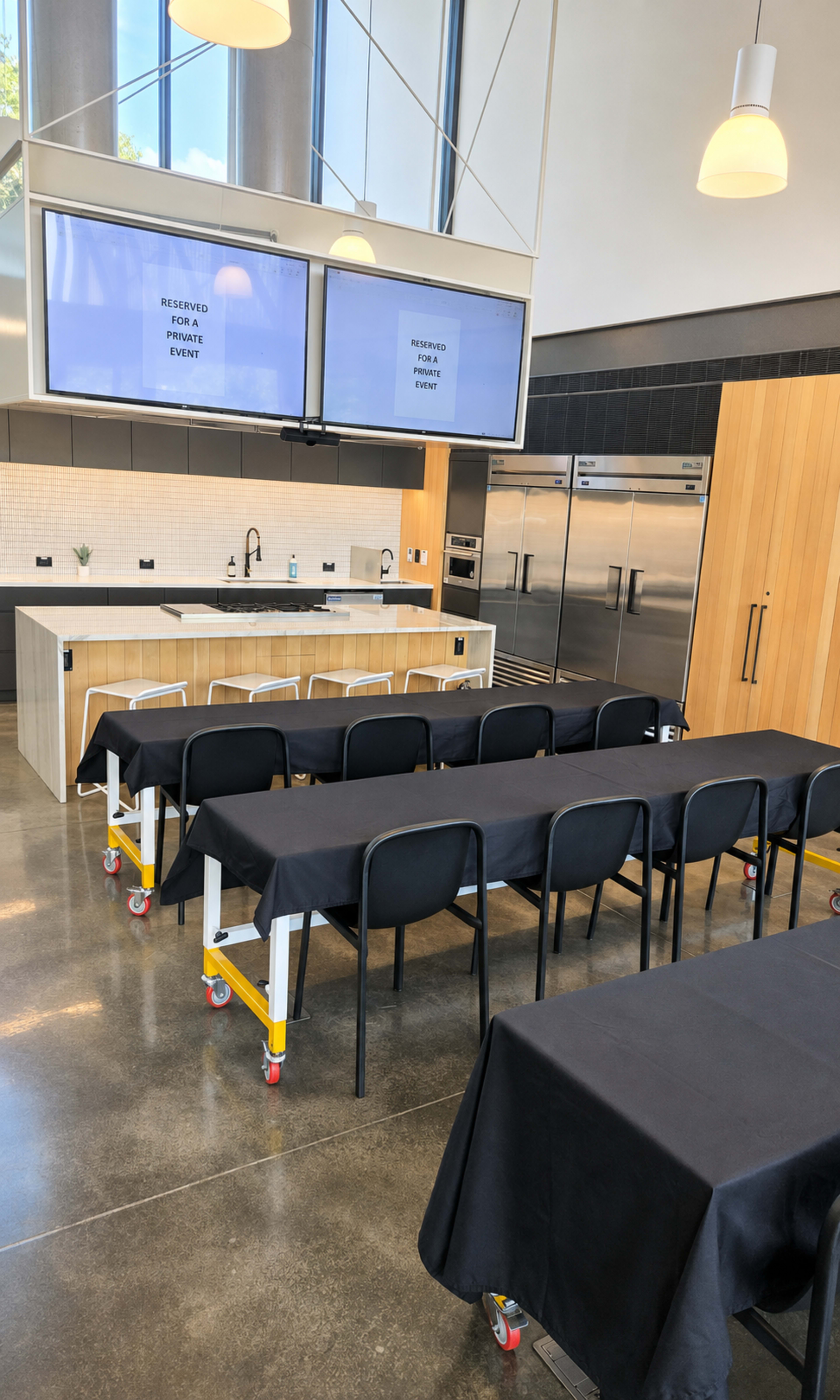 The image shows a modern event space with rows of black-covered tables, a kitchen area in the background, and two large screens displaying "RESERVED FOR PRIVATE EVENT."