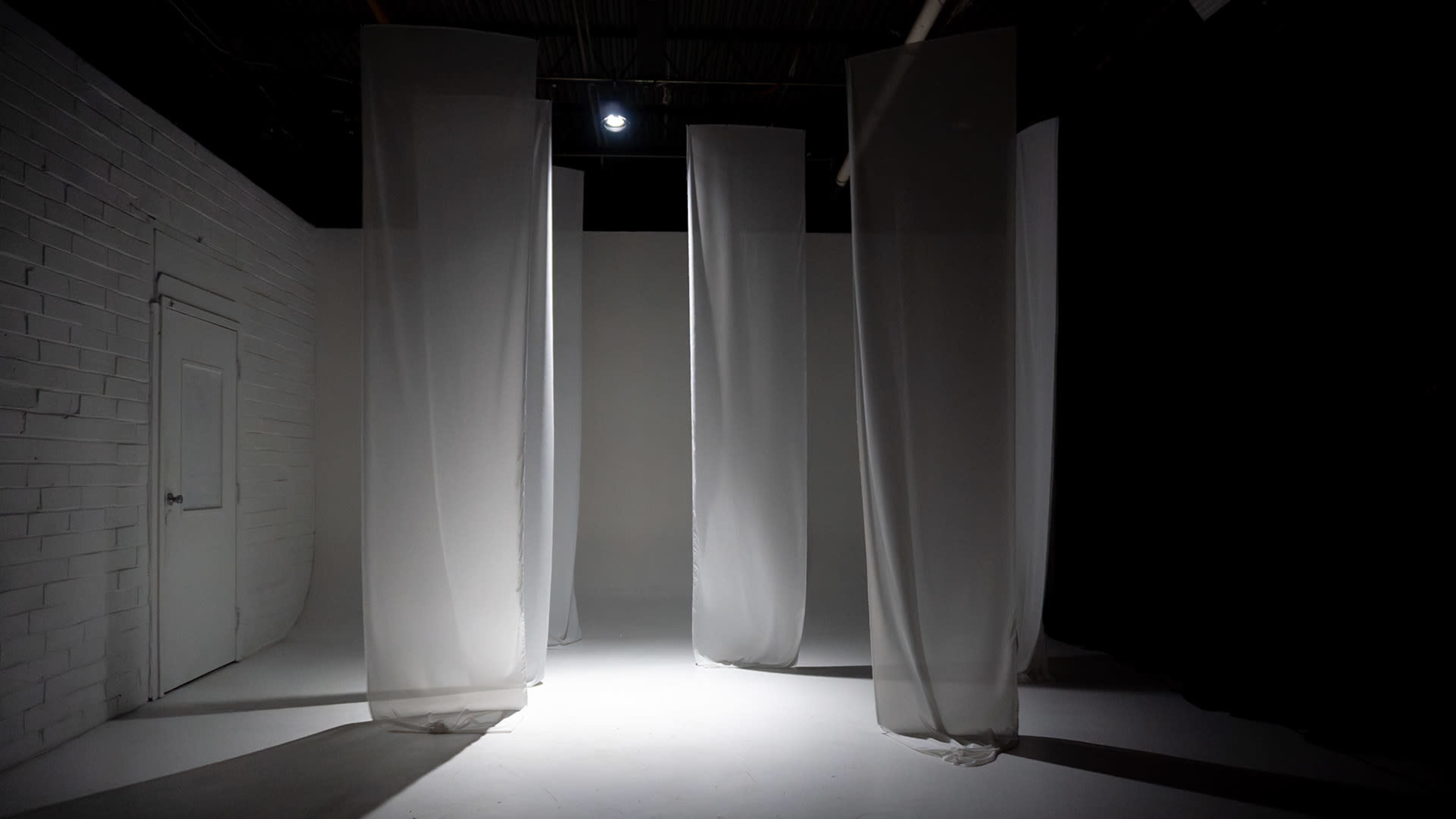 The image shows a dimly lit room with white fabric panels hanging from the ceiling amid a stark white floor and a white brick wall.