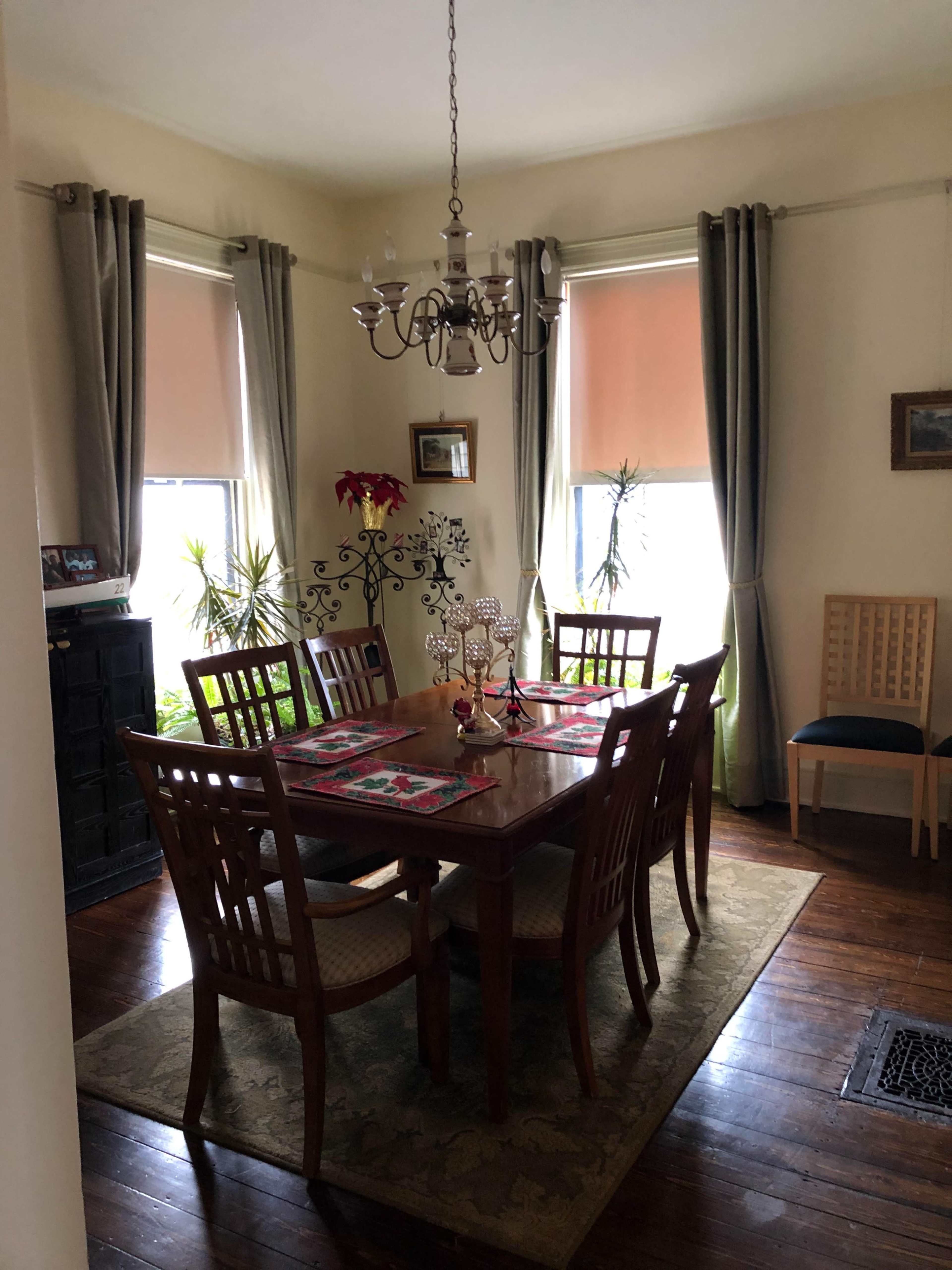 A dining room features a wooden table with eight chairs, a chandelier overhead, and natural light coming through two windows adorned with curtains.