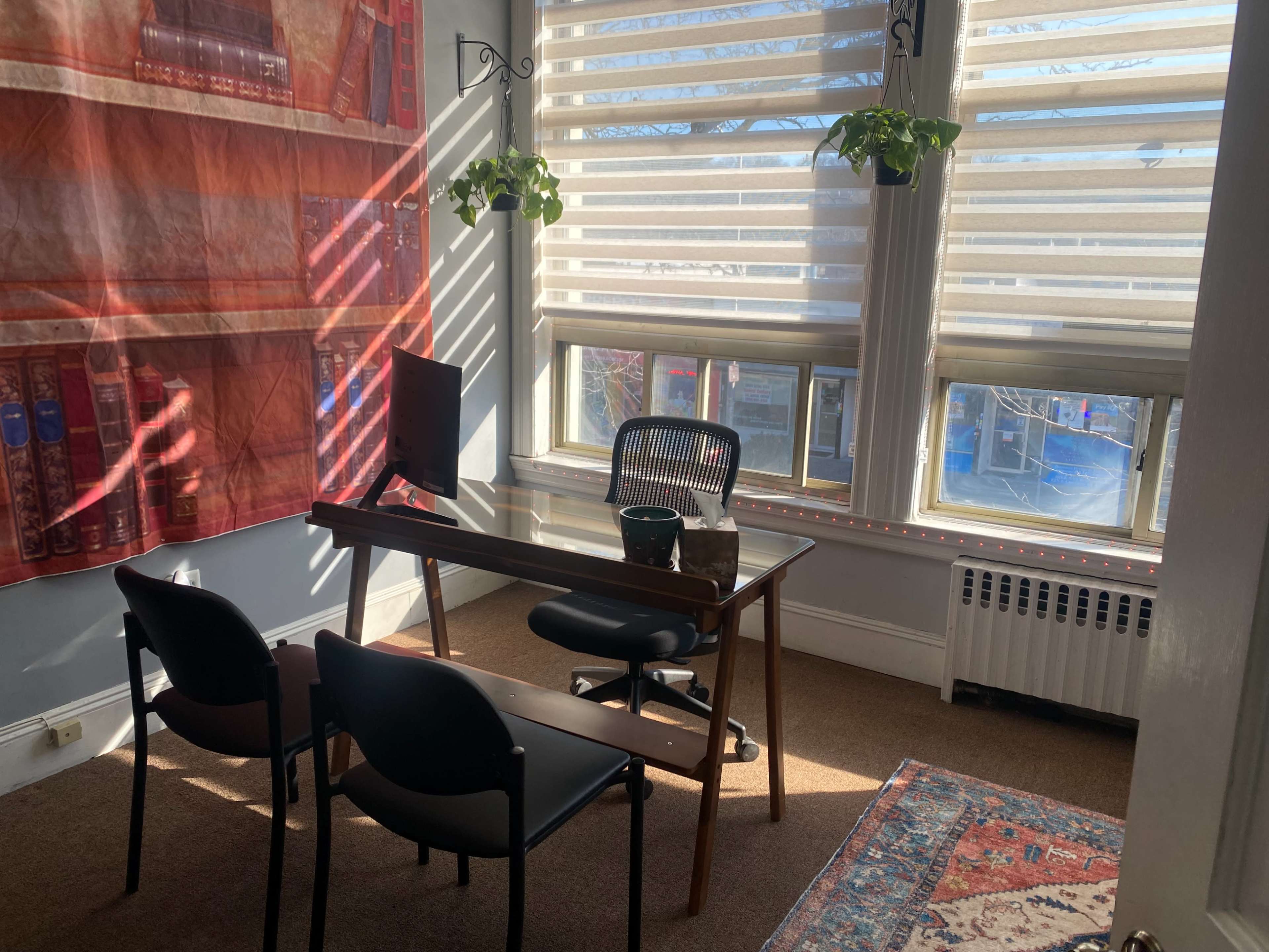 A small office space features a wooden desk, an office chair, two black chairs, and large windows with blinds letting in sunlight.
