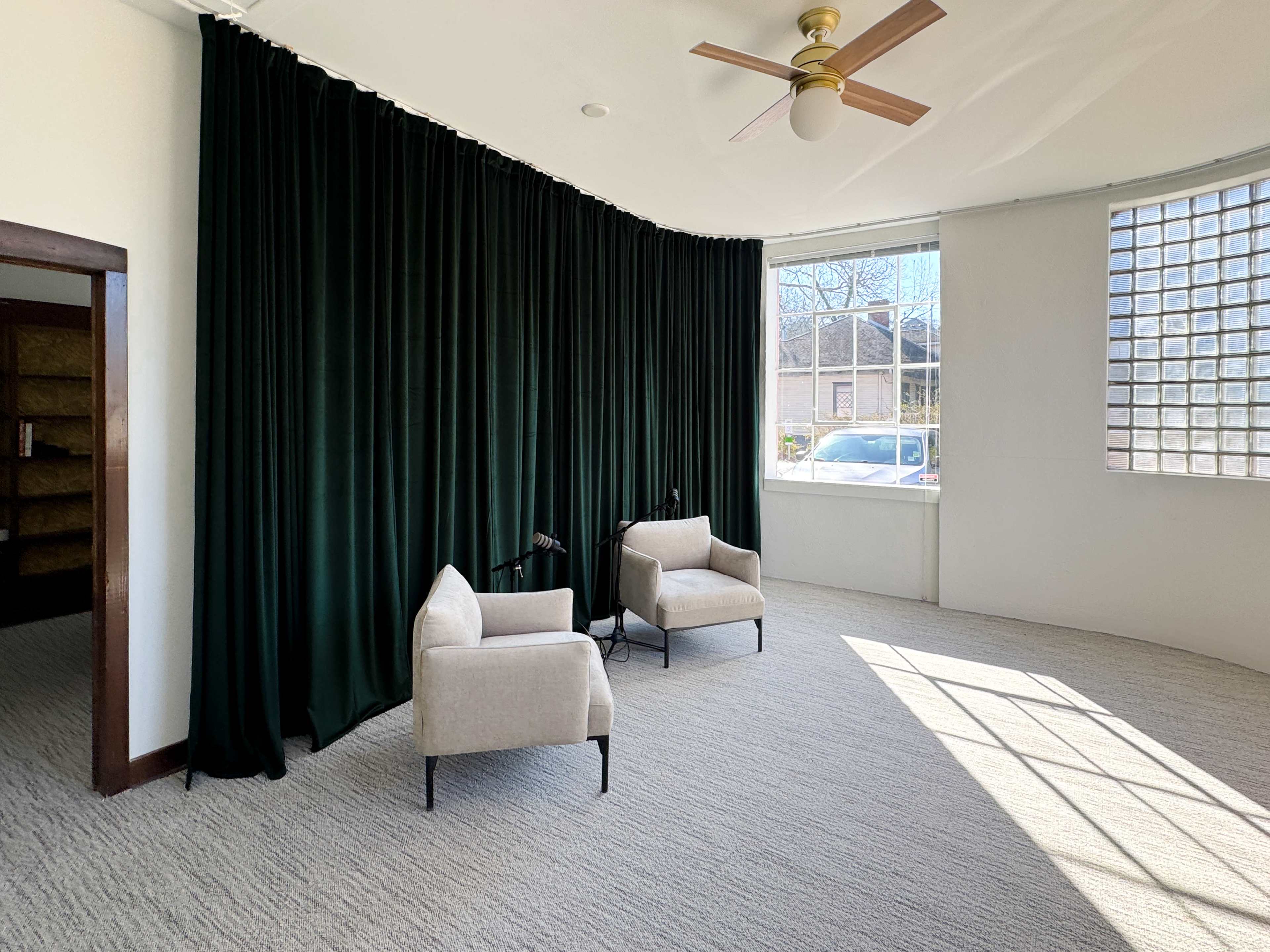 The image shows a bright room featuring two light-colored chairs next to a large window with green curtains and a ceiling fan.