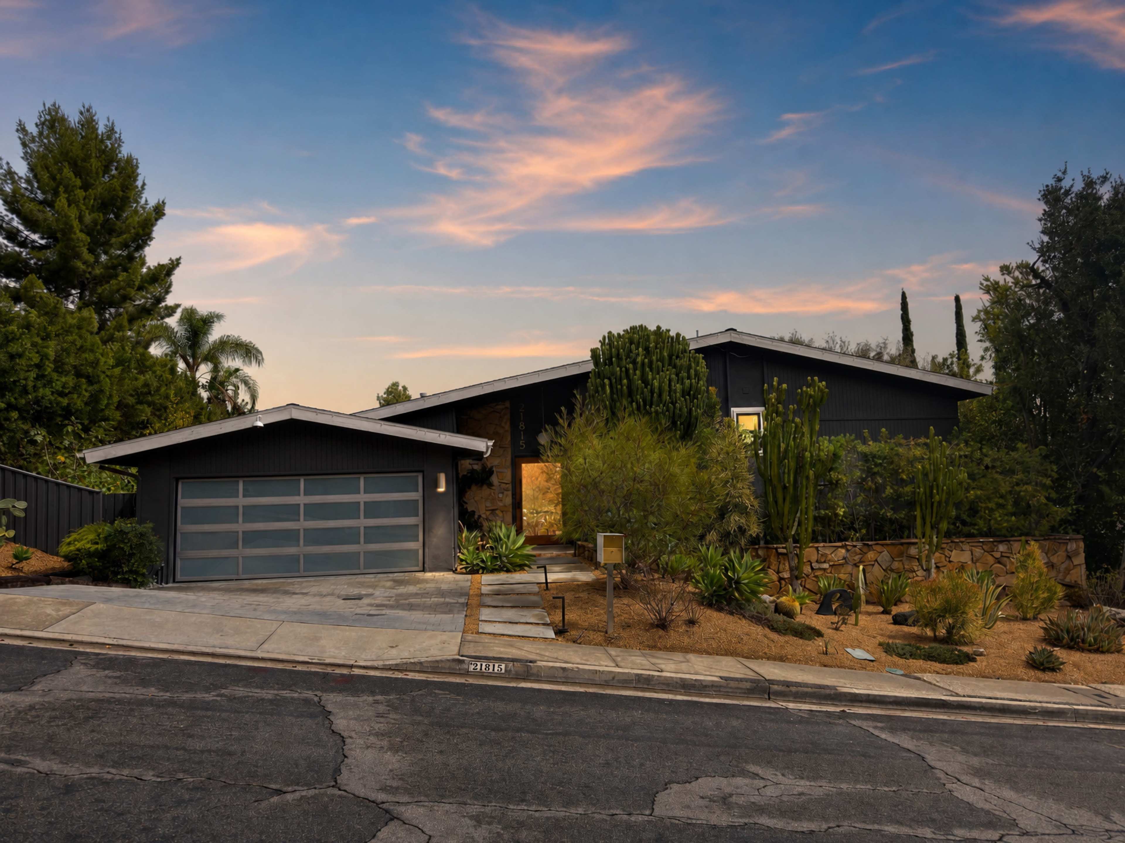 The image shows a modern, single-story house with a dark exterior, a garage, and landscaped greenery surrounding it, set against a twilight sky.