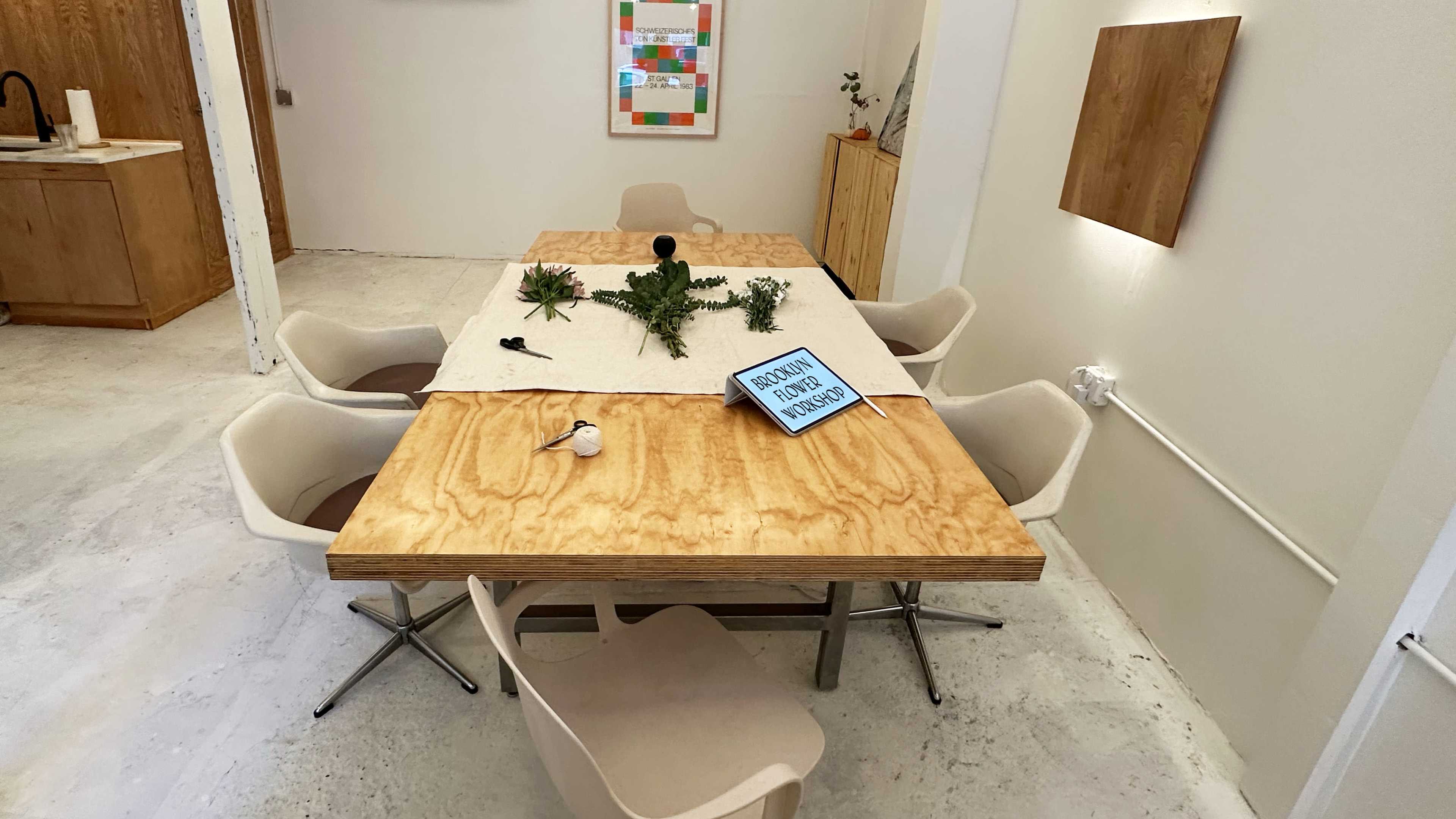 A wooden table with a beige cloth on top is surrounded by six chairs, with various plants and a sign displayed on the table in a well-lit room.