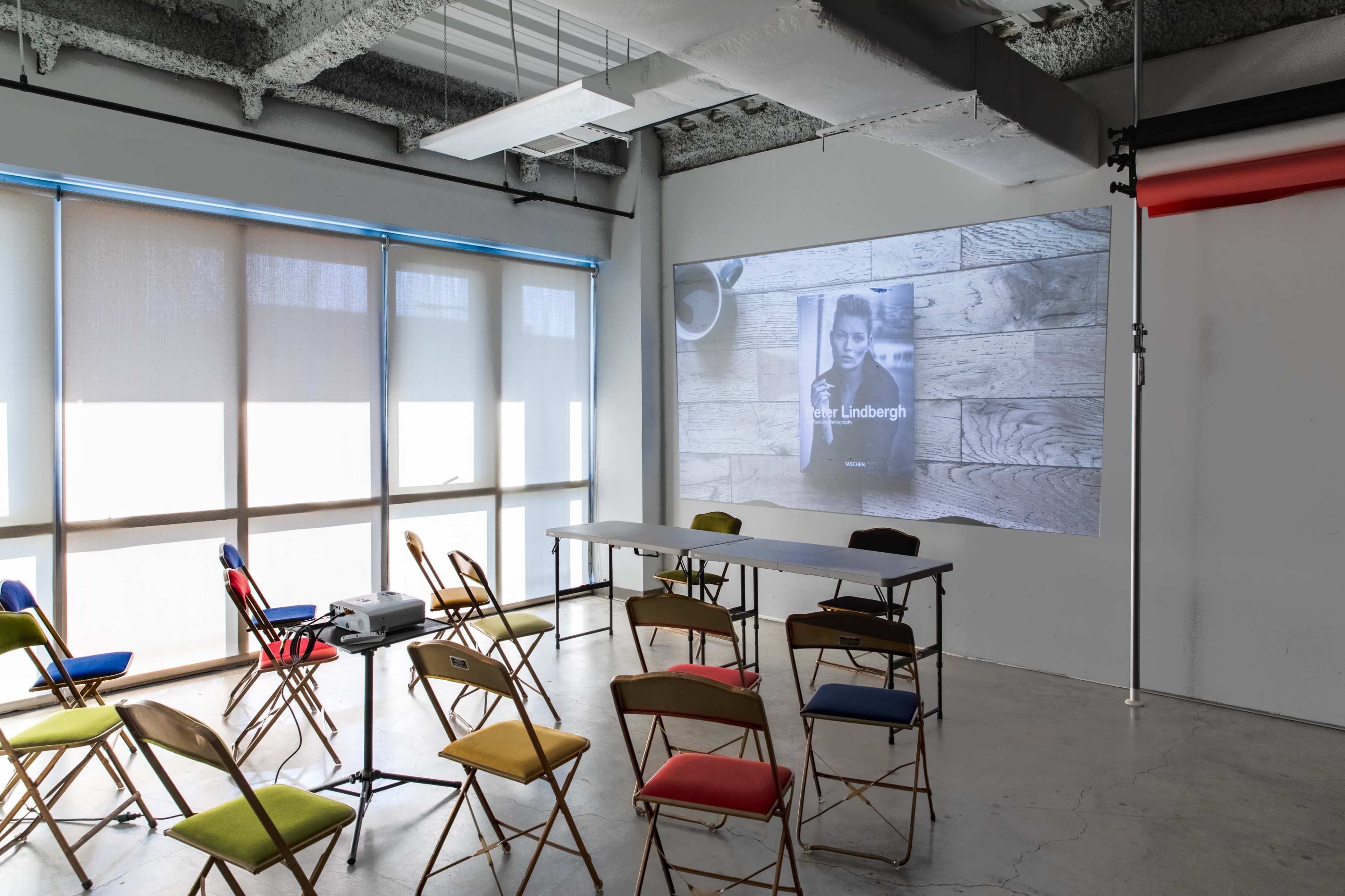 A projector displays an image on a wall in a room set up with rows of colorful chairs and a long table.