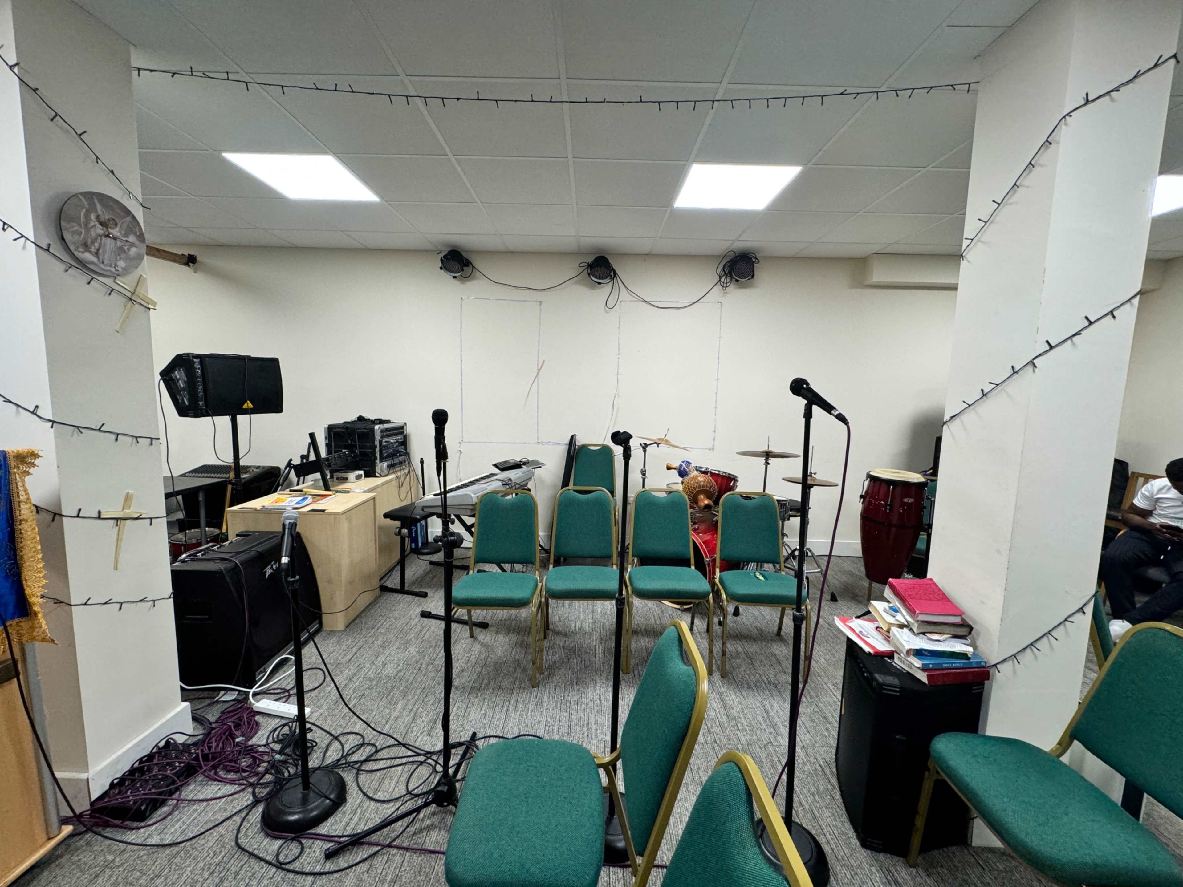 A room set up for a music practice session, featuring empty green chairs arranged facing a microphone setup near music equipment and instruments.