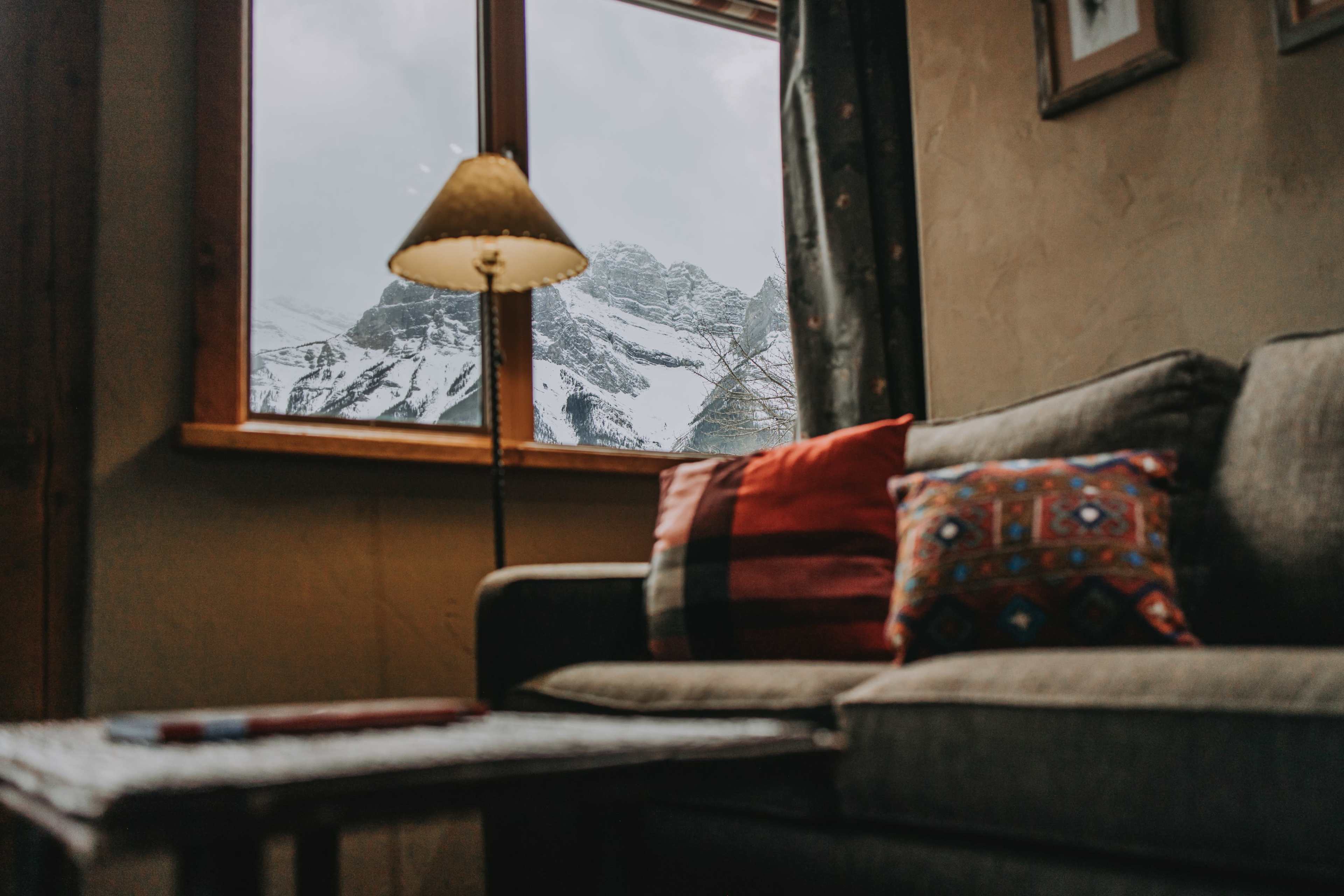 A cozy living room features a gray sofa with decorative pillows, a side table, and a lamp, alongside a large window showcasing snowy mountains in the background.