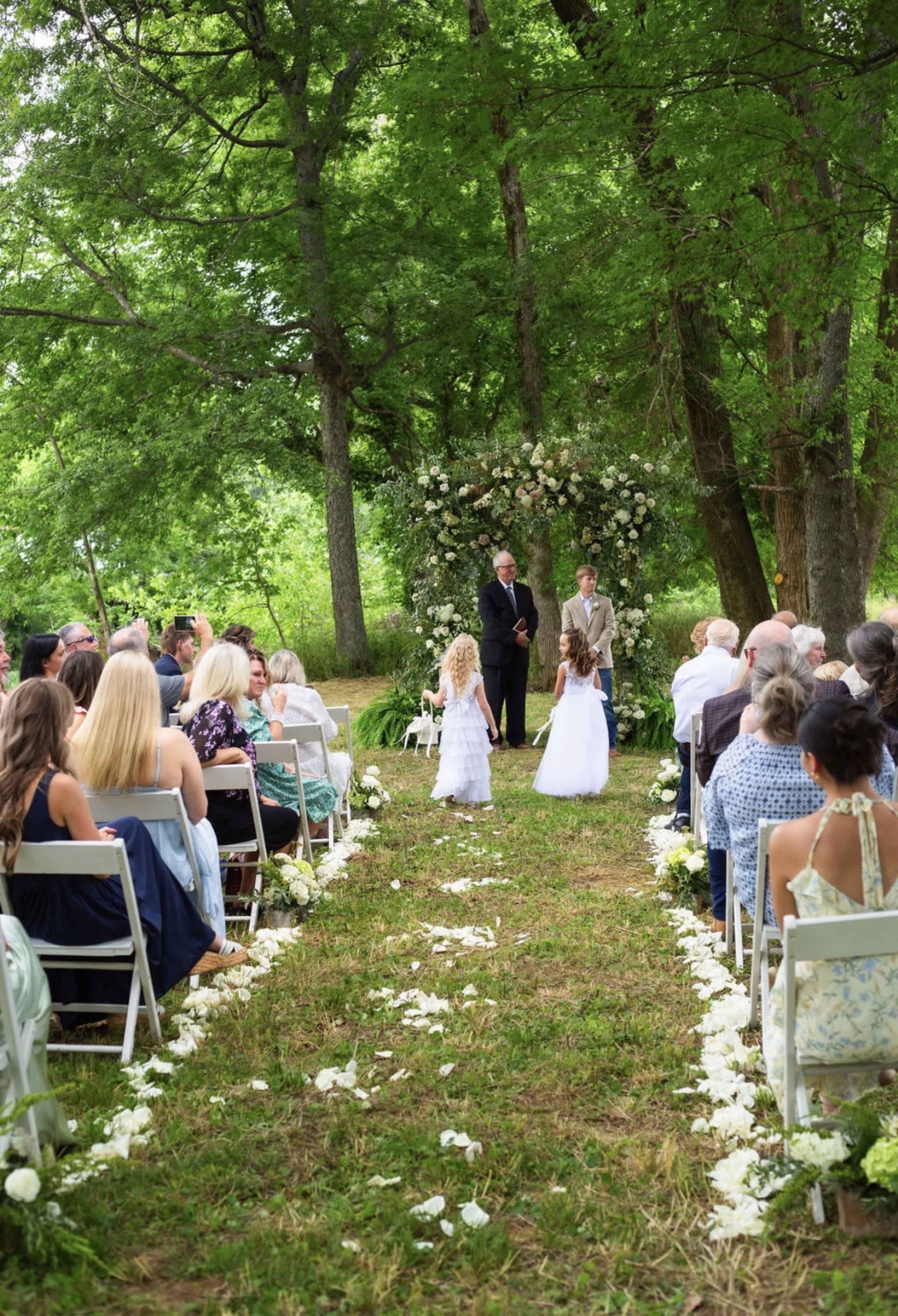A wedding ceremony takes place outdoors under a floral arch, with guests seated on either side of a flower-strewn aisle.