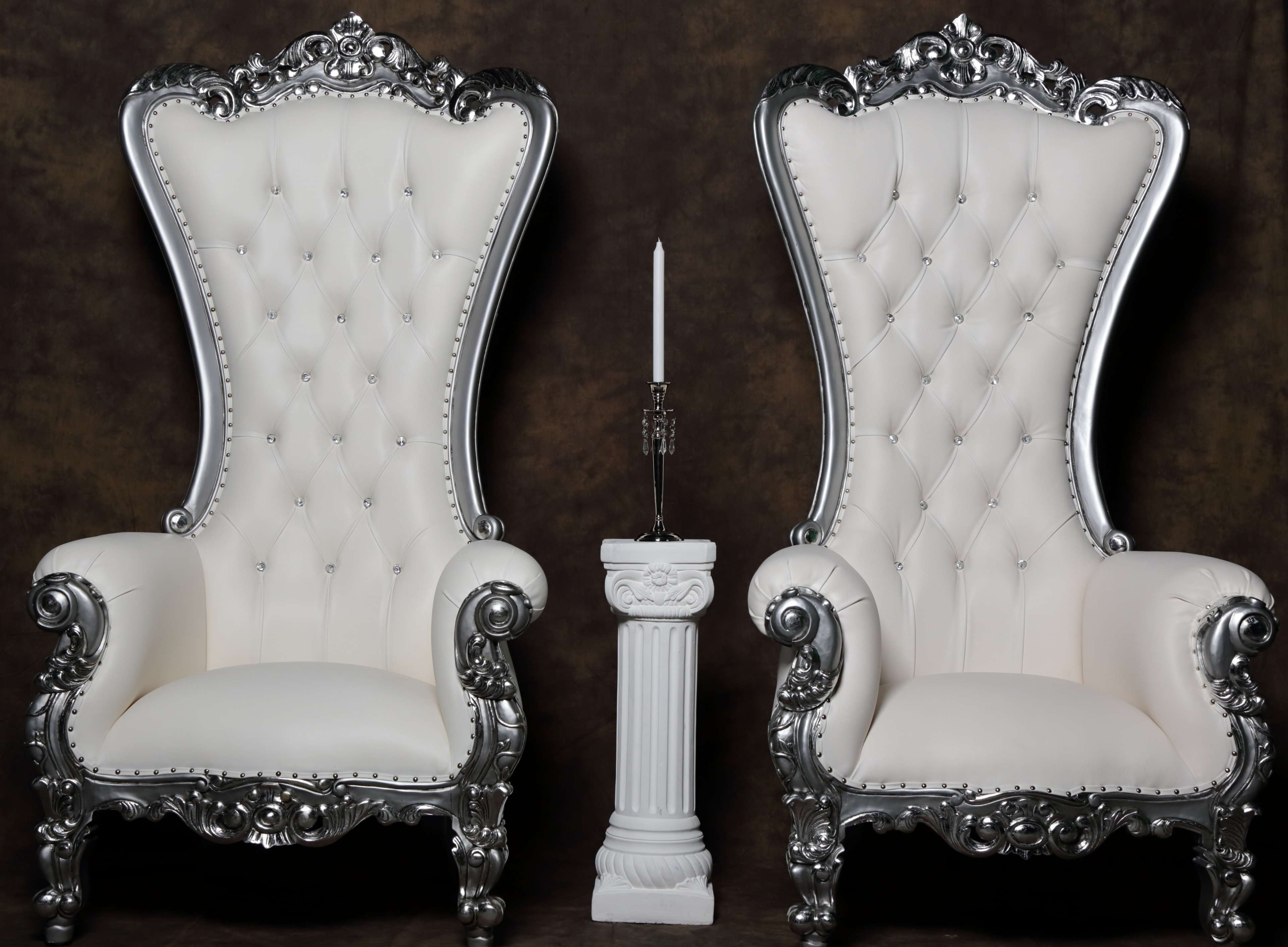 Two ornate white and silver throne chairs positioned on either side of a small white column with a candle on it.