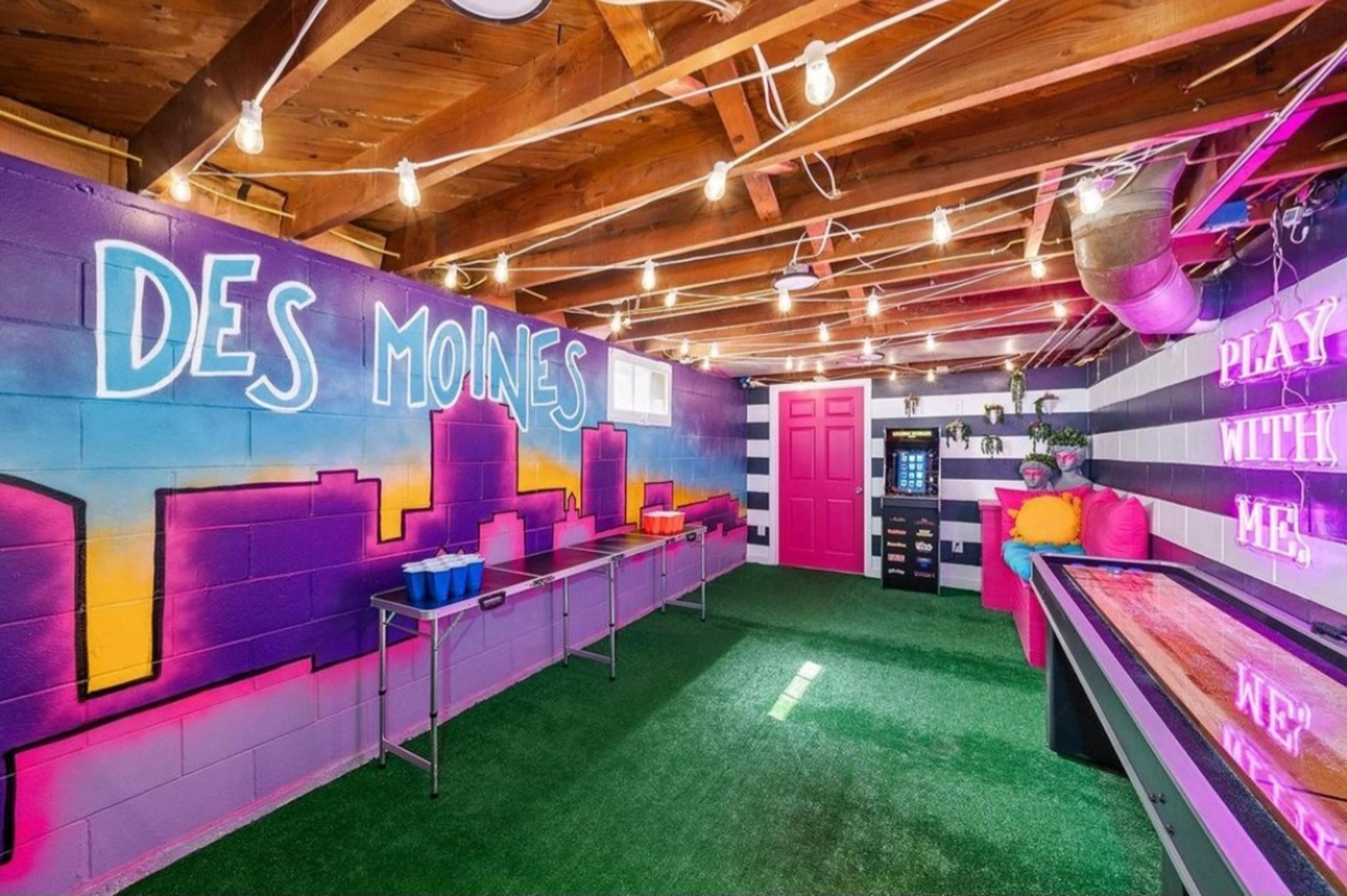 A brightly colored indoor space with purple and pink walls, artificial grass flooring, and string lights, featuring tables and a vending machine.