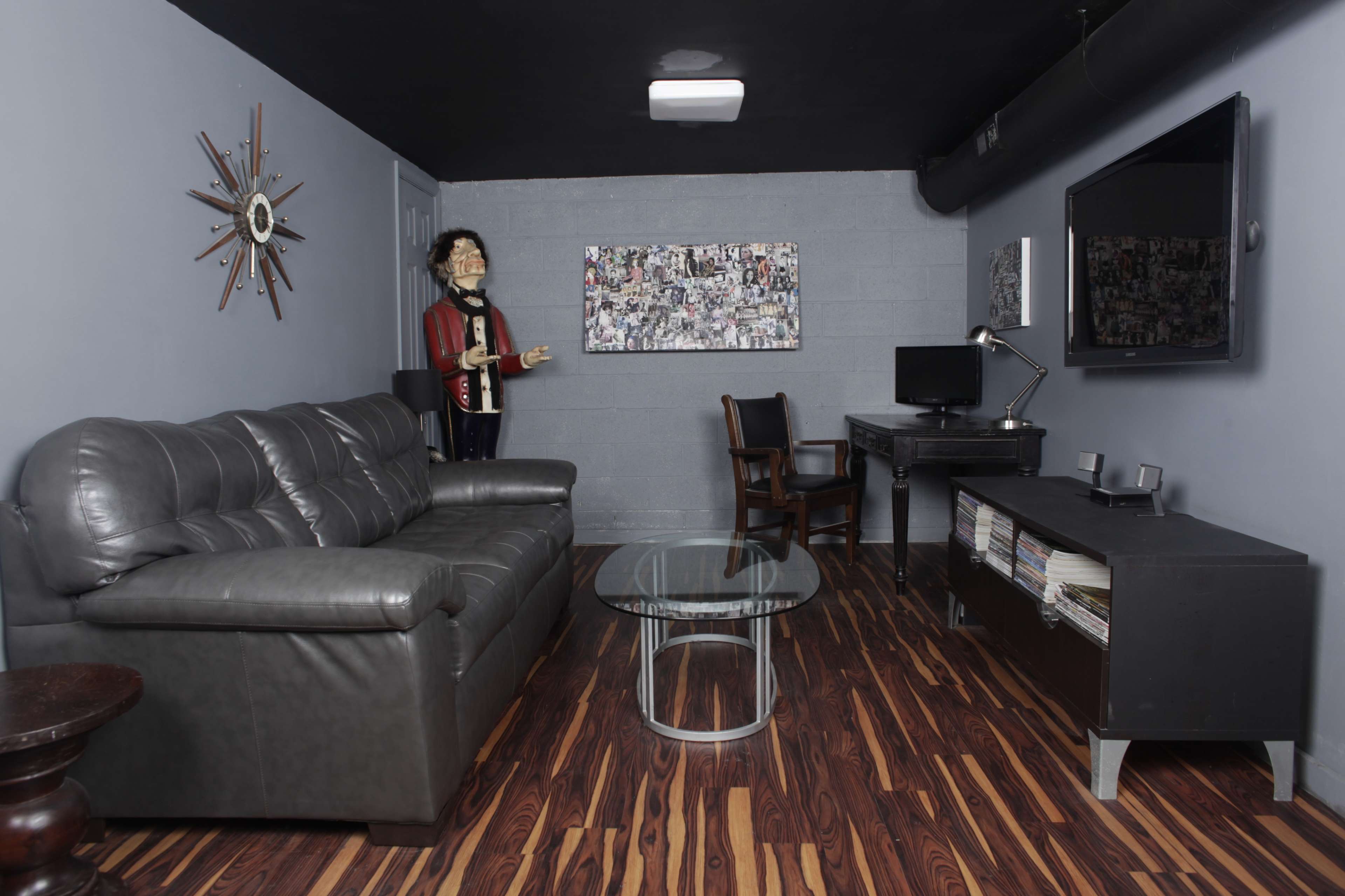 The room features a gray wall, a black leather couch, a glass coffee table, a wooden chair, a desk with a computer, and a life-sized mannequin dressed in a suit.