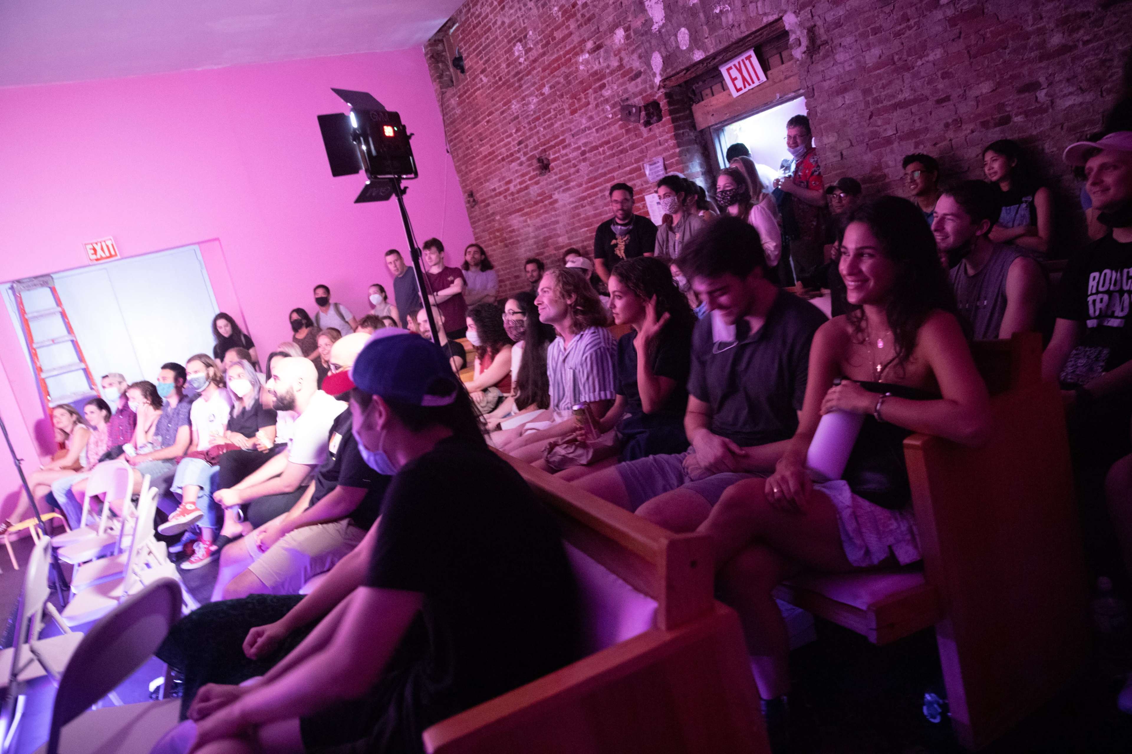 A diverse audience sits in a brightly colored room, attentively watching a performance onstage.