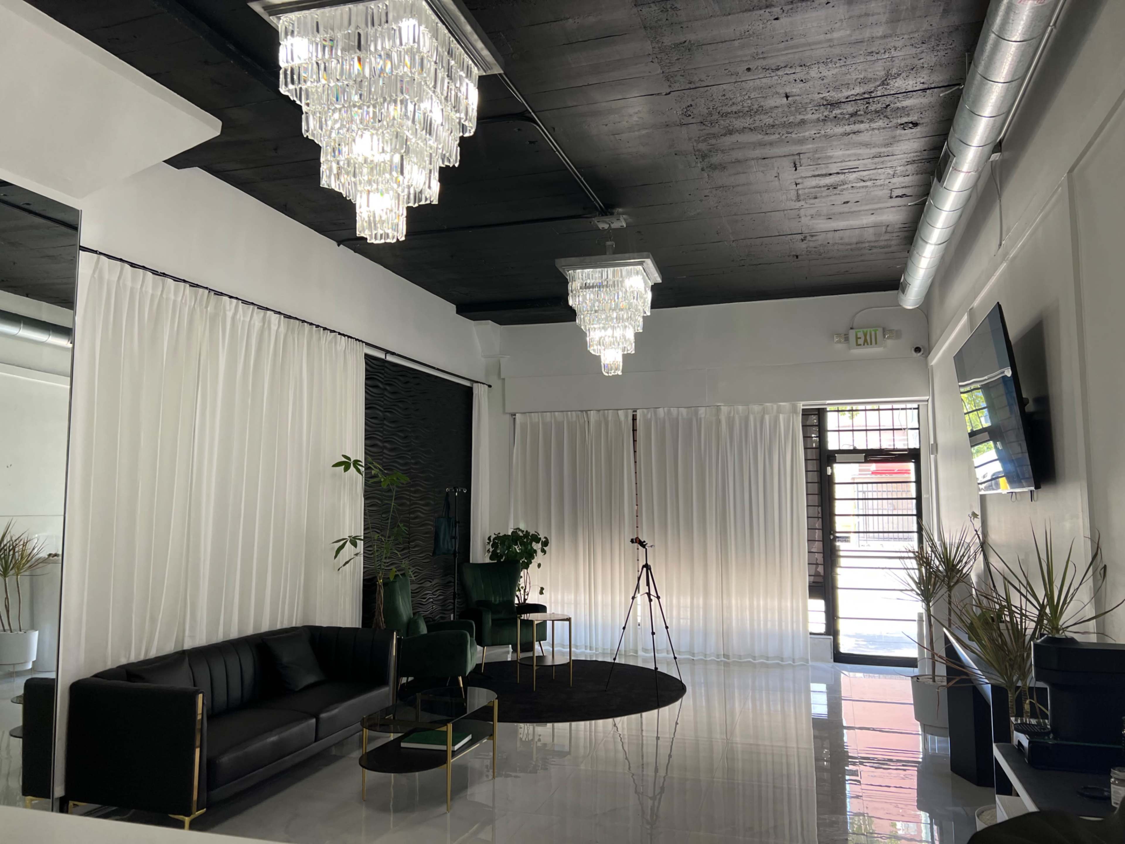 The image features a sleek, modern interior with two crystal chandeliers, a black leather sofa, a round black rug, and large windows with white curtains.
