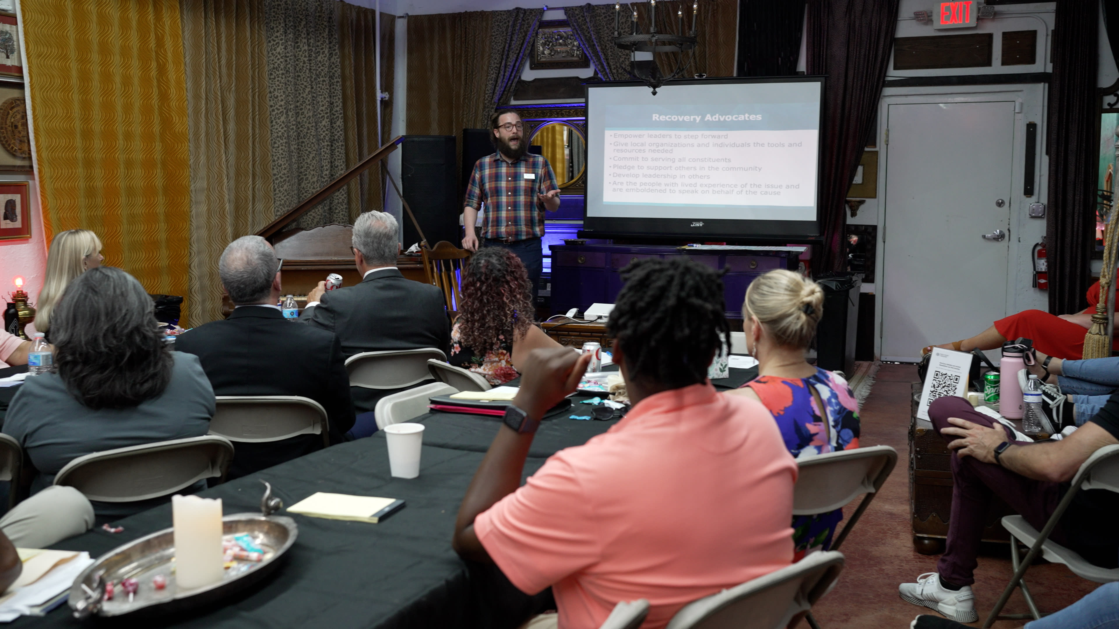A speaker presents information about recovery advocates to an audience seated at tables in a conference setting.