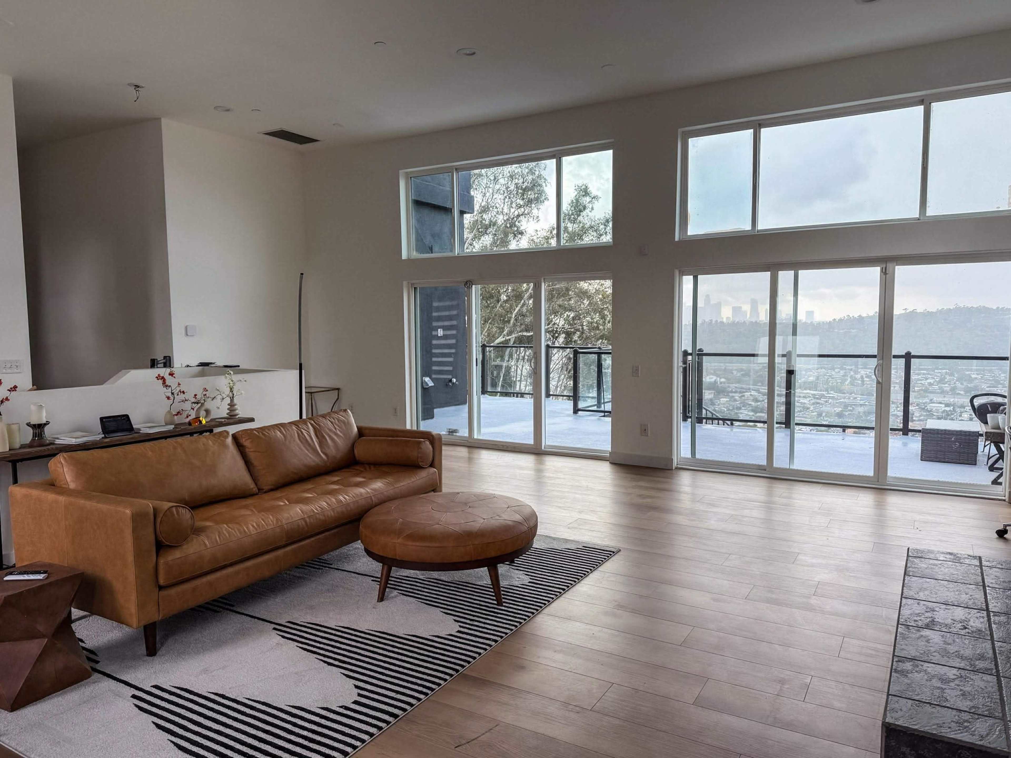 The image shows a modern living room with a brown leather sofa, an ottoman, and large windows that overlook a cityscape.