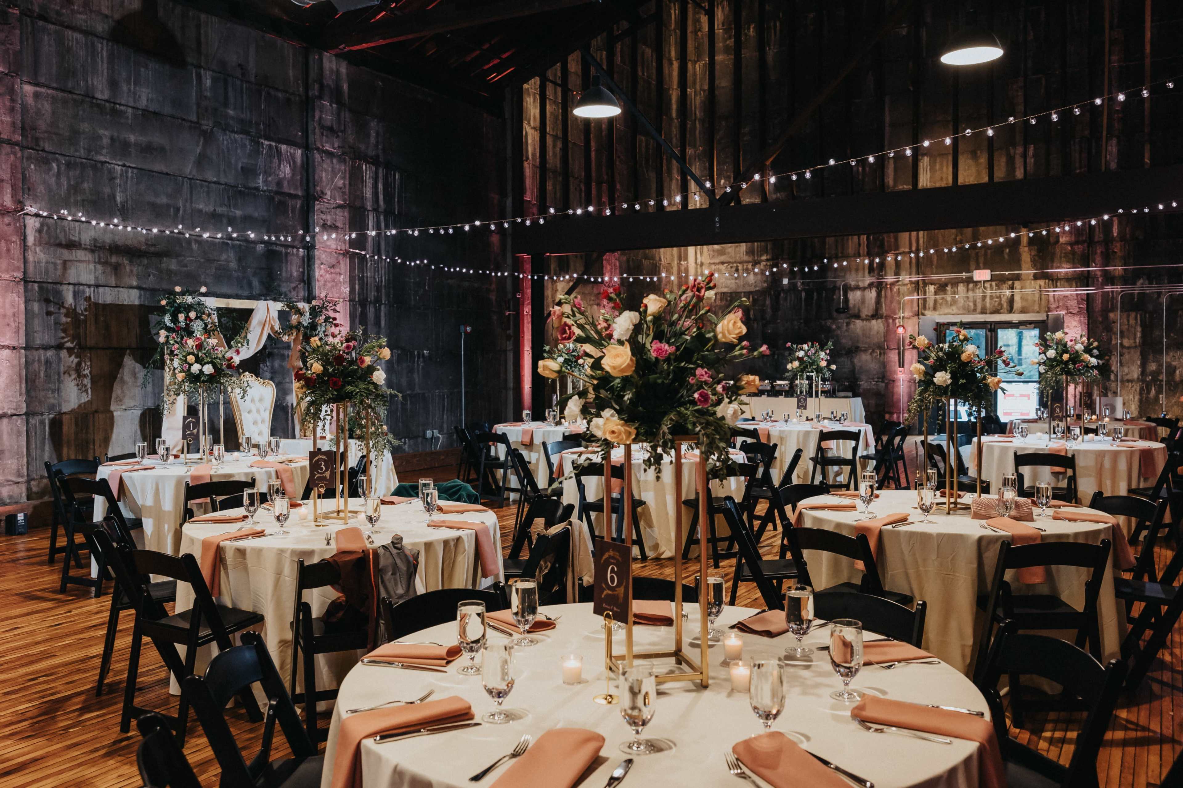 A large event space features round tables elegantly set with floral centerpieces, soft pink napkins, and rustic decor, illuminated by string lights overhead.
