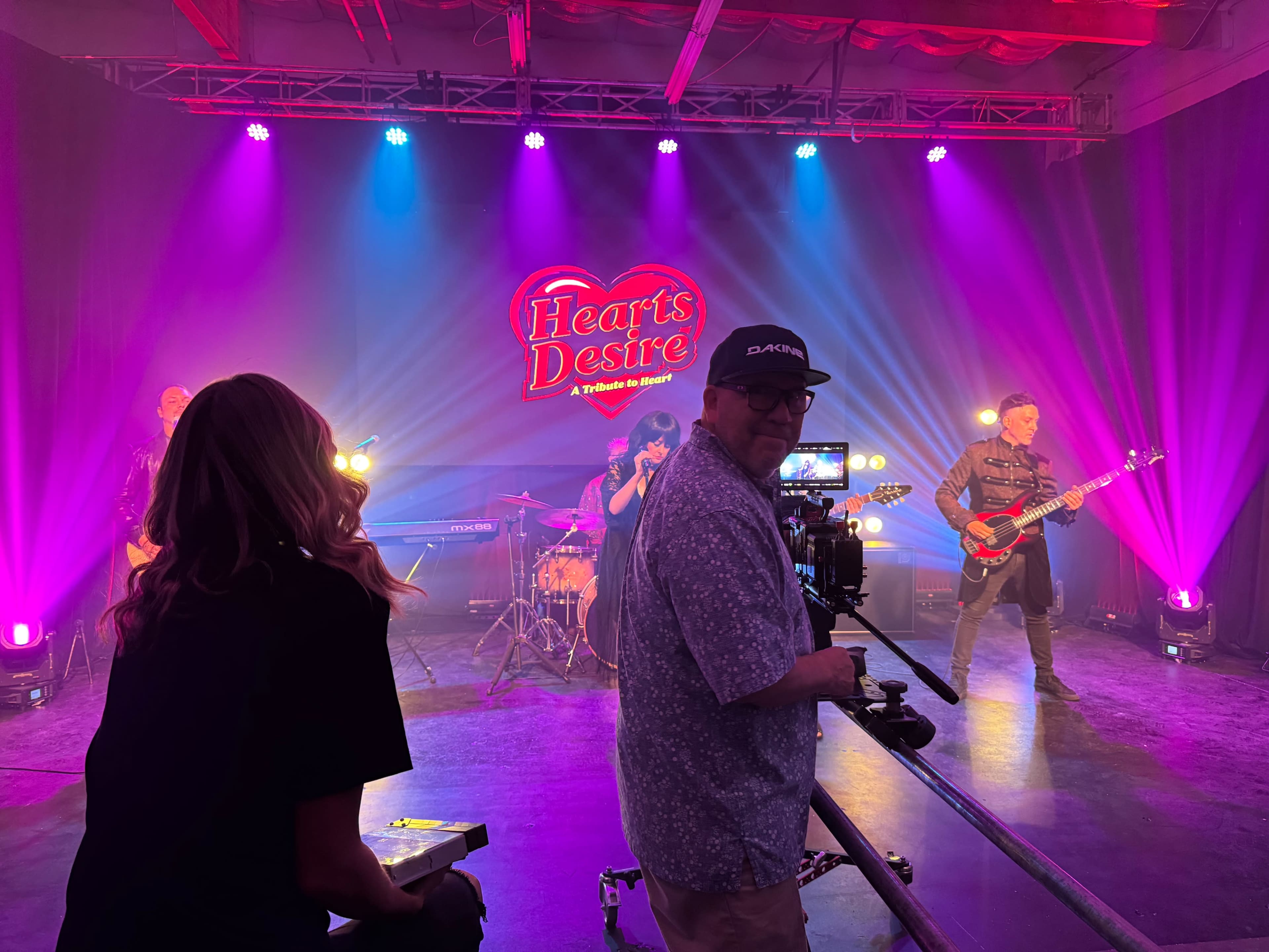 A band performs onstage with colorful lights and a backdrop featuring the title "Hearts Desire."