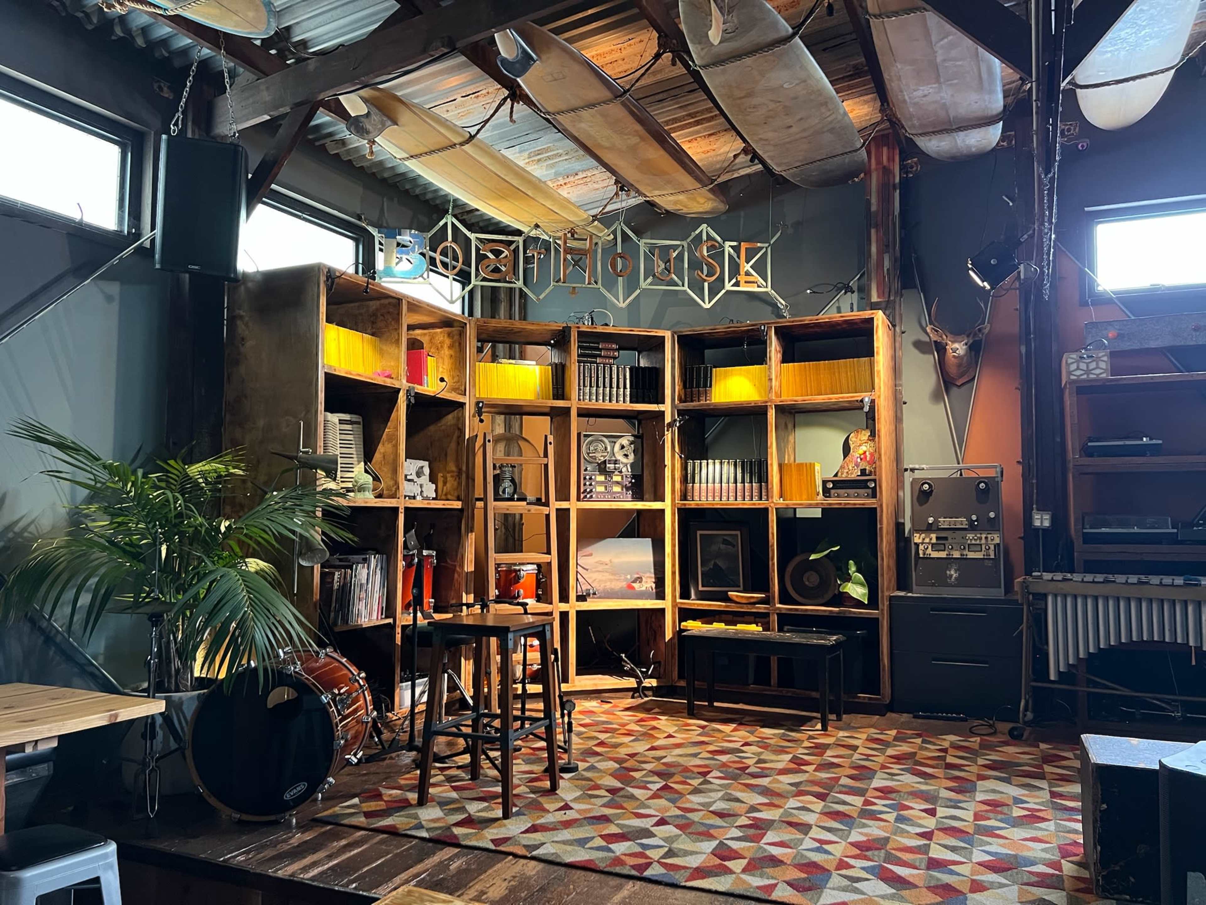The image shows a cozy interior space with a colorful rug, wooden shelves filled with books and decorations, a drum set, and a sign that reads "Gearhouse."
