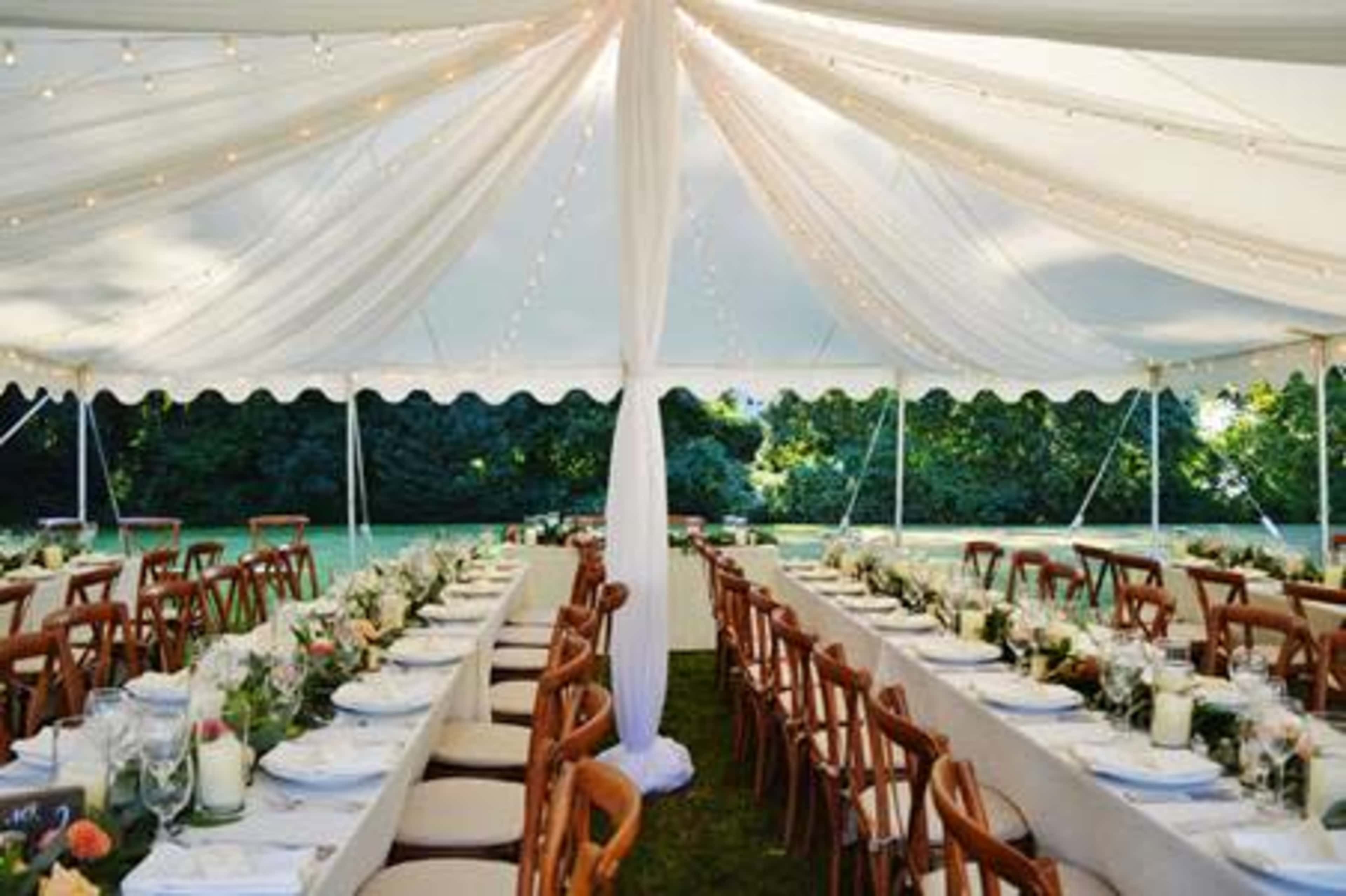 A large outdoor tent is set up with long tables adorned with white tablecloths, flowers, and candles for a gathering or event.