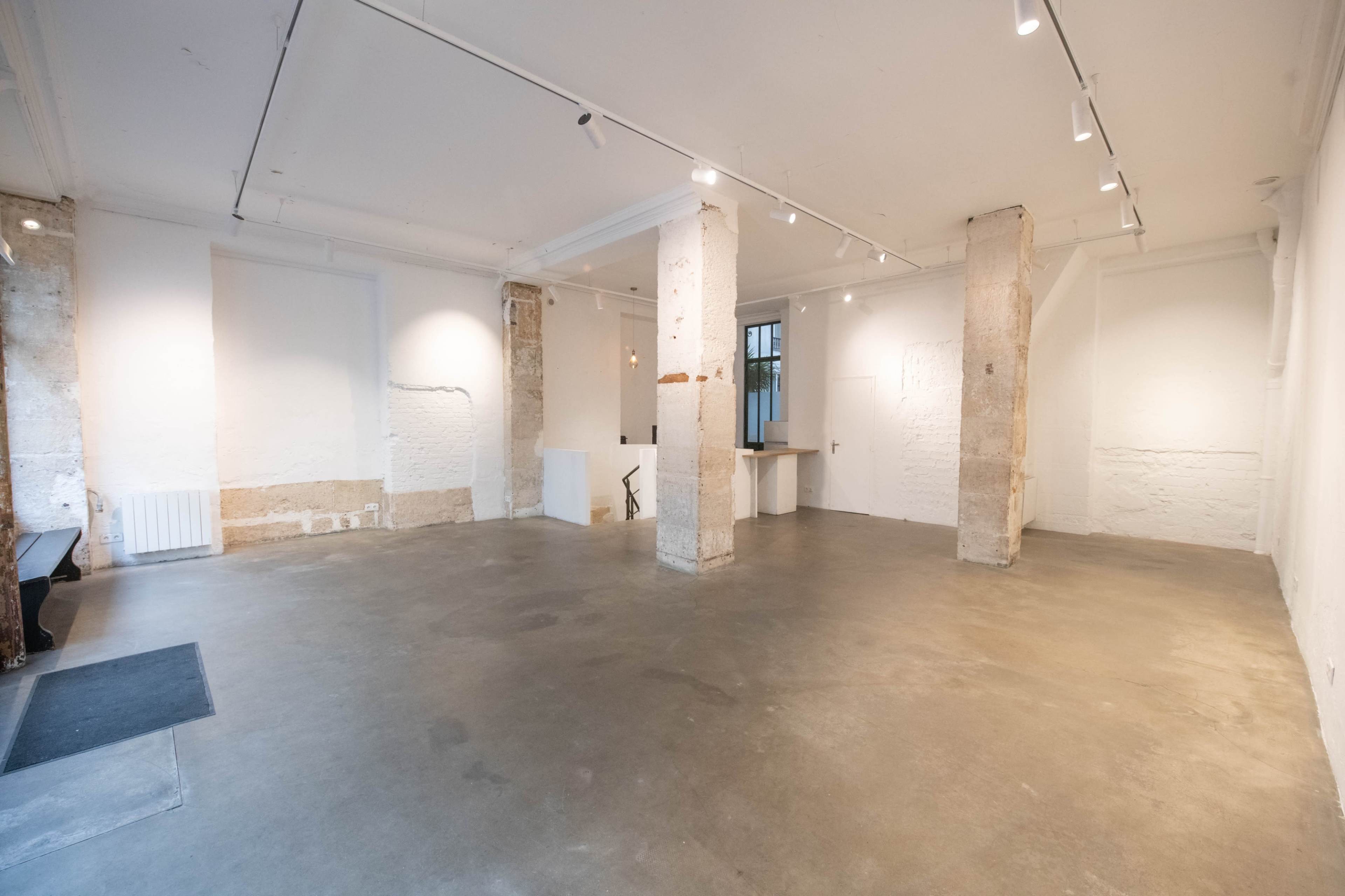 A spacious, minimalist gallery interior with concrete floors and exposed pillars, featuring white walls and soft overhead lighting.