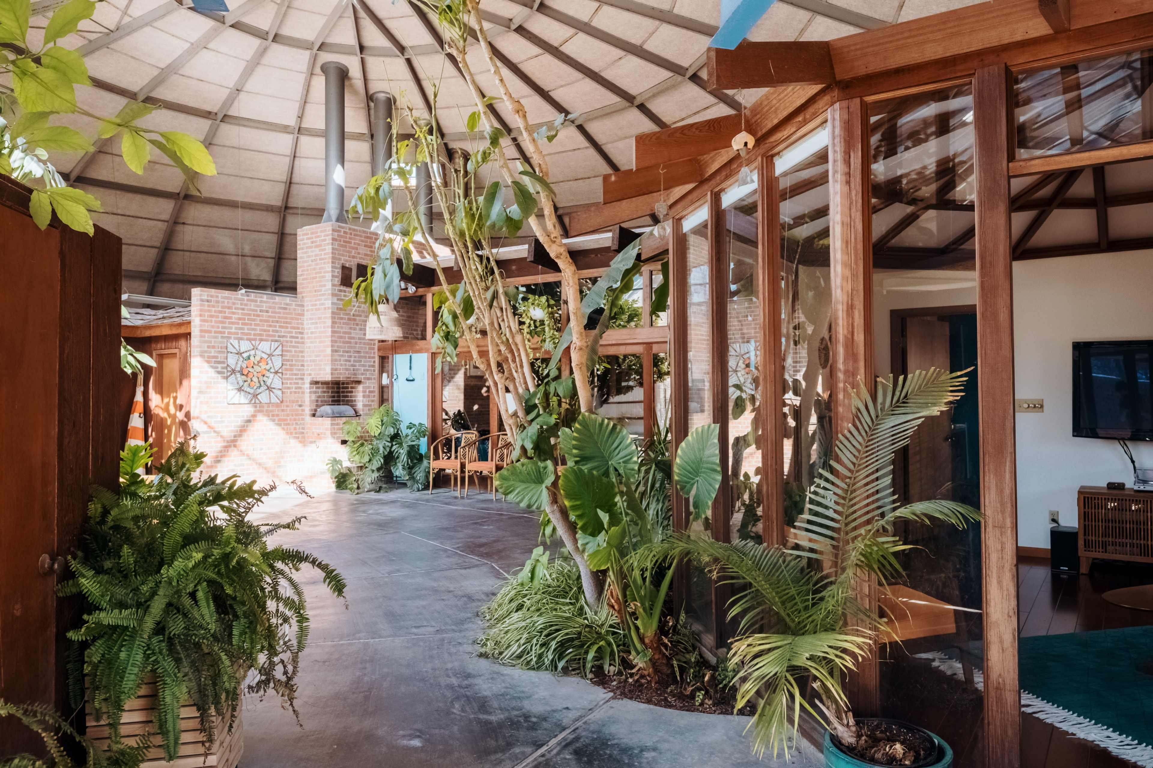 The image shows a circular interior space with wooden beams, large glass windows, and various plants integrated into the design.