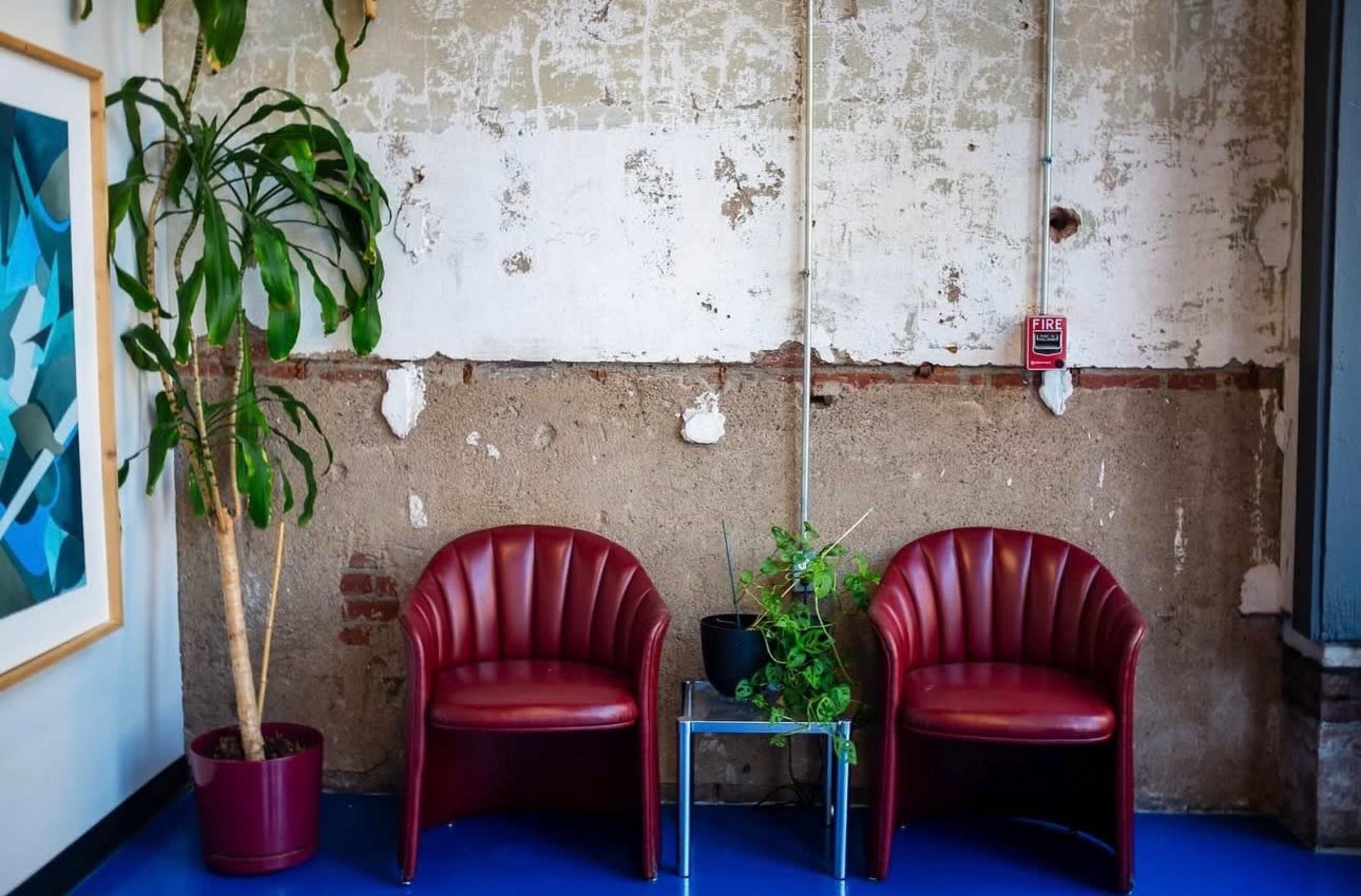 Two red chairs are positioned against a weathered wall with exposed bricks, accompanied by a small plant in a pot and a glass table.