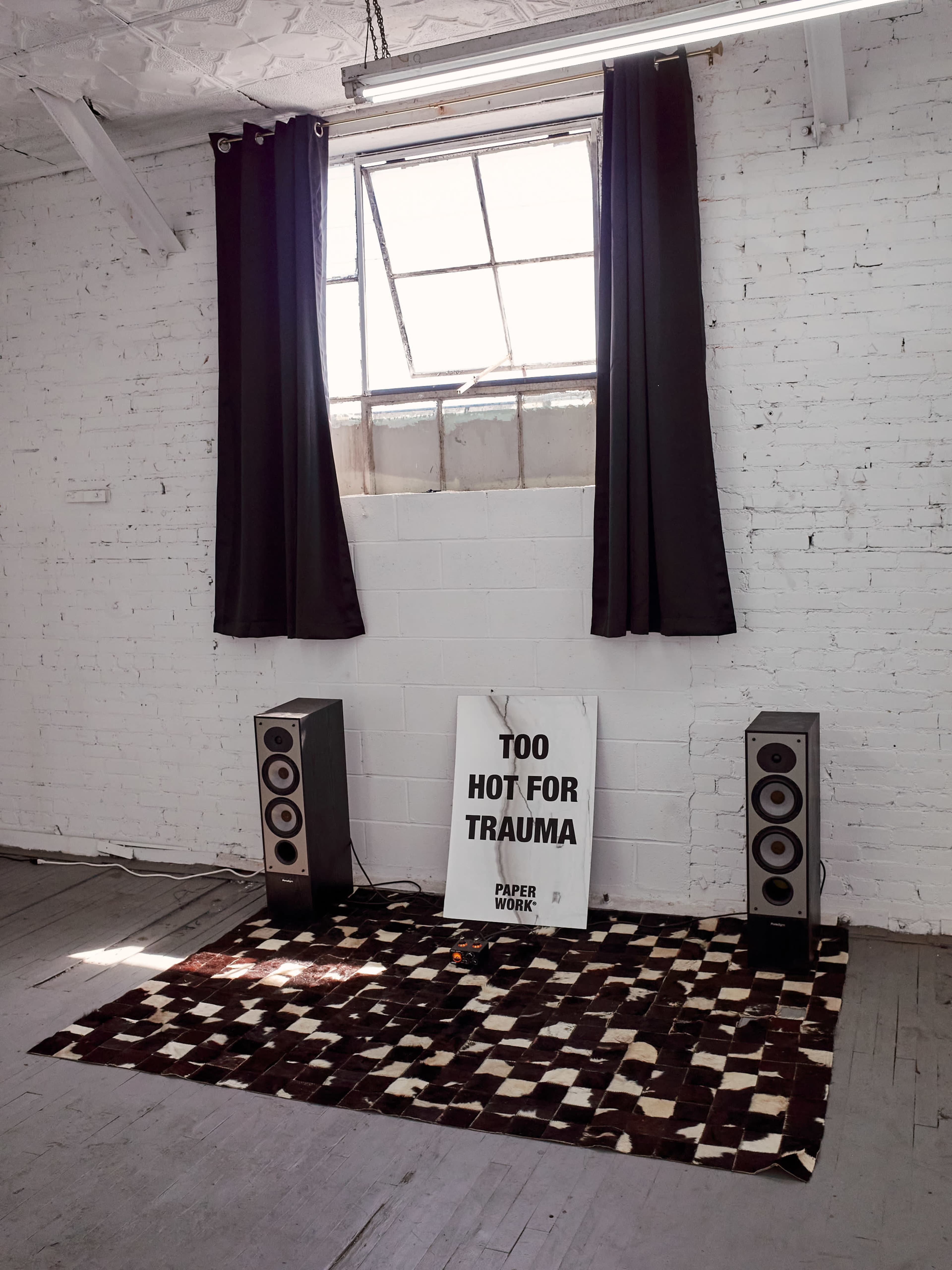 The image shows a minimalist room with two tall speakers flanking a large sign that reads "TOO HOT FOR TRAUMA," positioned on a patterned rug beneath a window with black curtains.