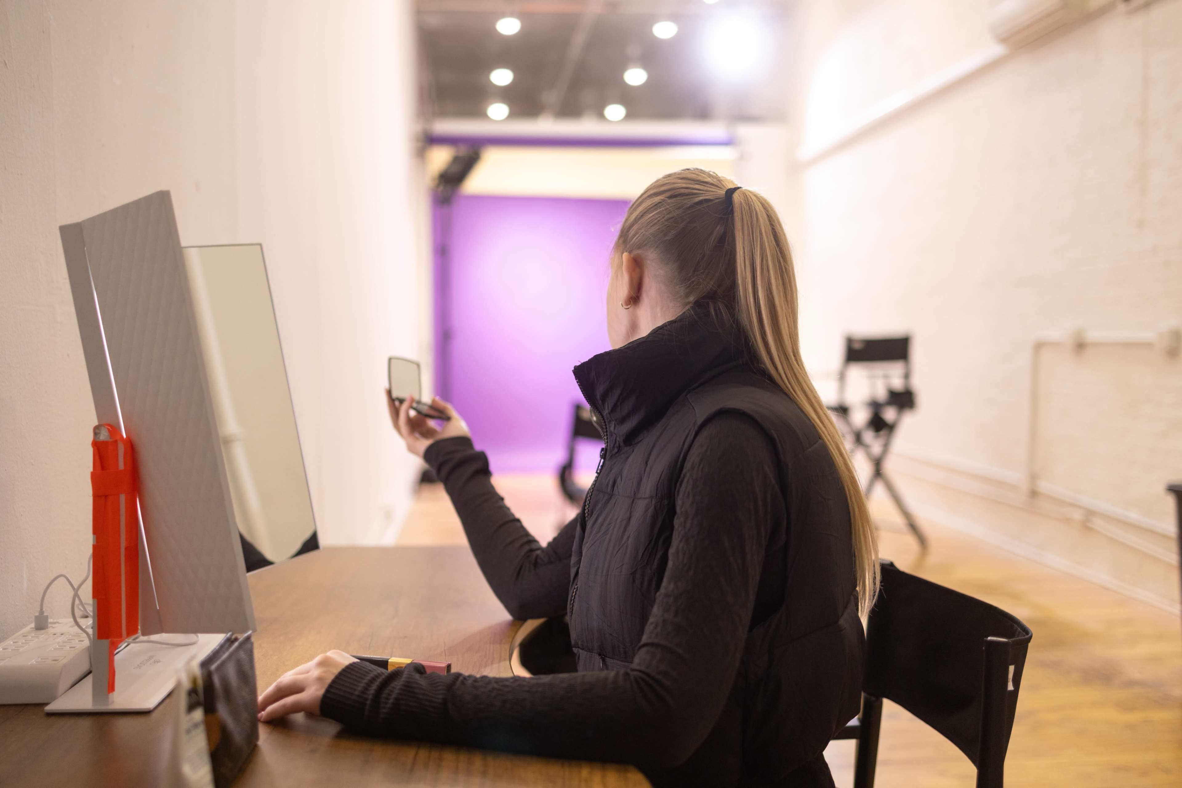 A person with a ponytail is sitting at a wooden table, holding a small mirror in a well-lit room with a purple backdrop.