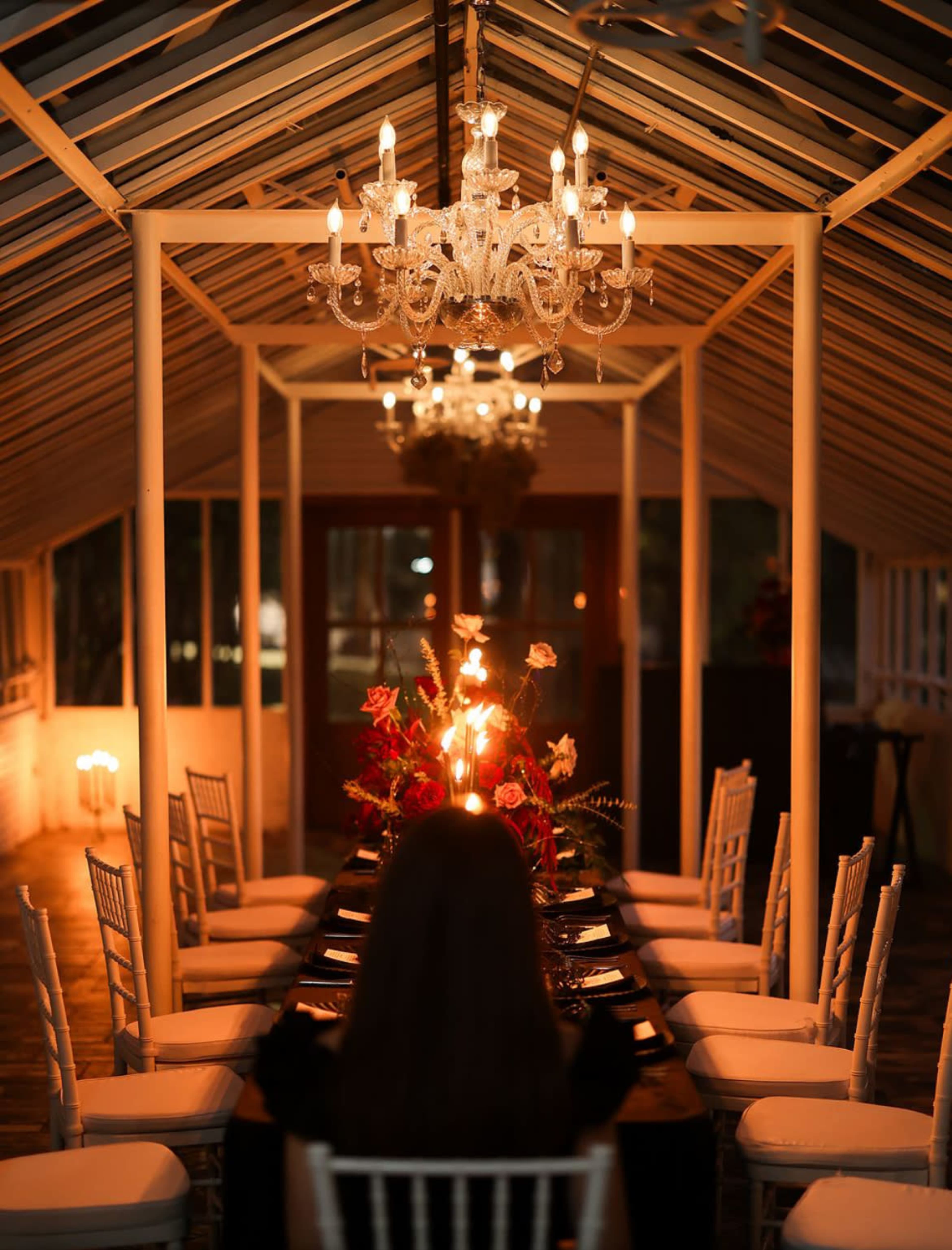 A long dining table is set in a glass greenhouse illuminated by chandeliers, with a person sitting at one end surrounded by decorated chairs.